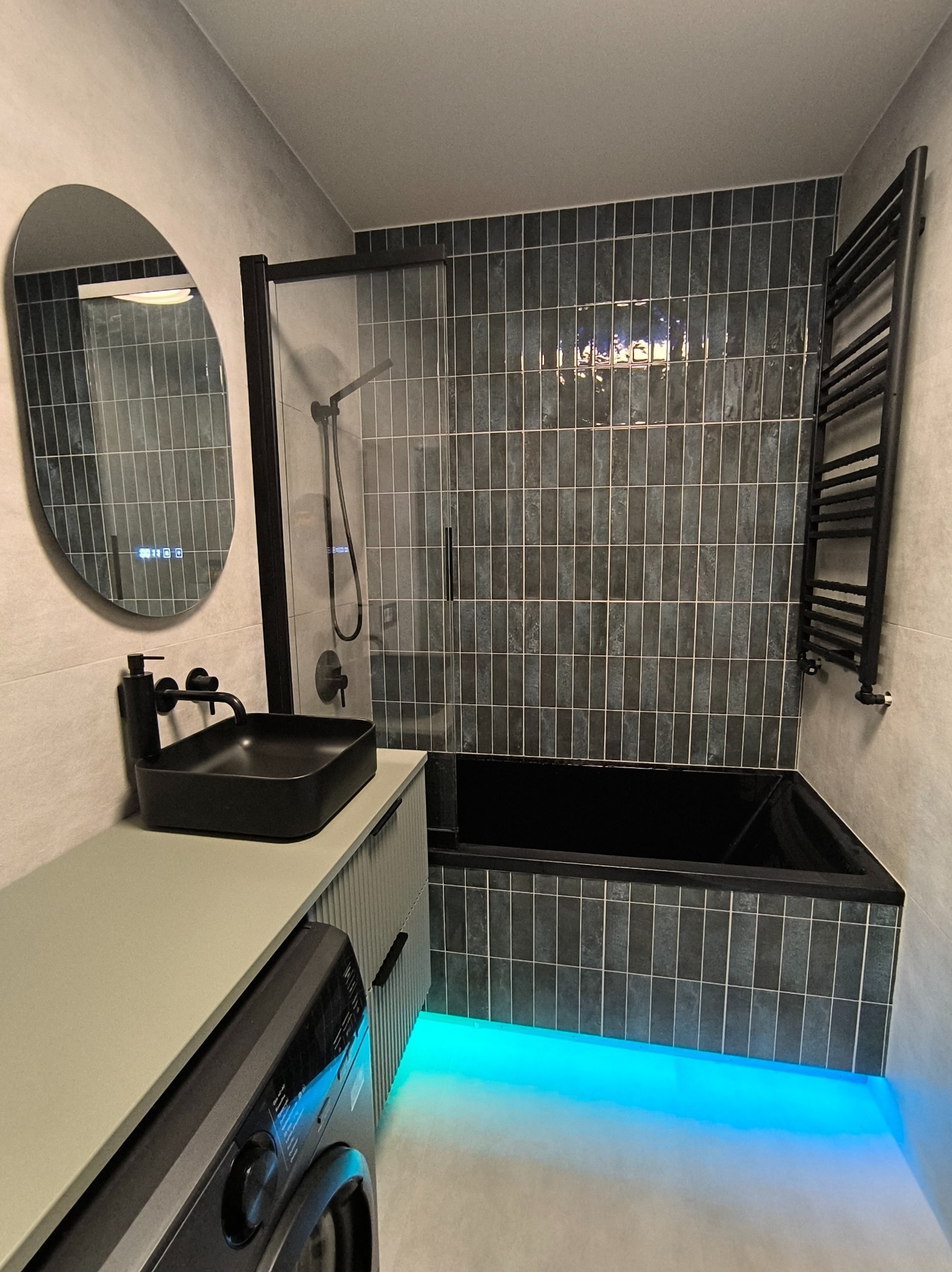 Nowoczesna łazienka z czarną armaturą, podświetlaną wanną i owalnym lustrem z wyświetlaczem. Ciemne płytki na ścianach i jasne na podłodze tworzą kontrast.