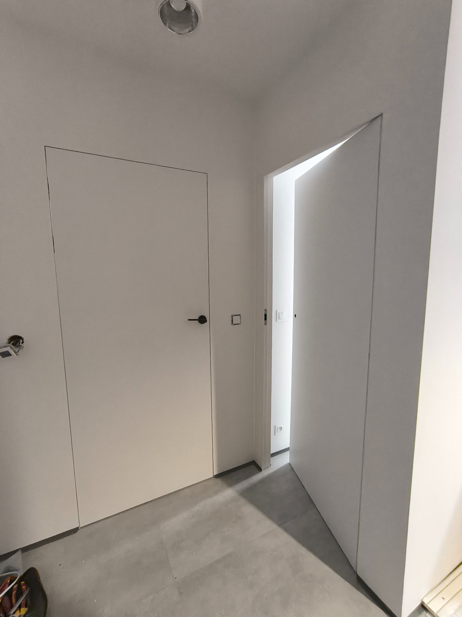 Minimalistyczne wnętrze: białe, ukryte drzwi z czarną klamką, szara podłoga, sufit z oświetleniem punktowym. Jedne drzwi otwarte, ukazują kolejne pomieszczenie.