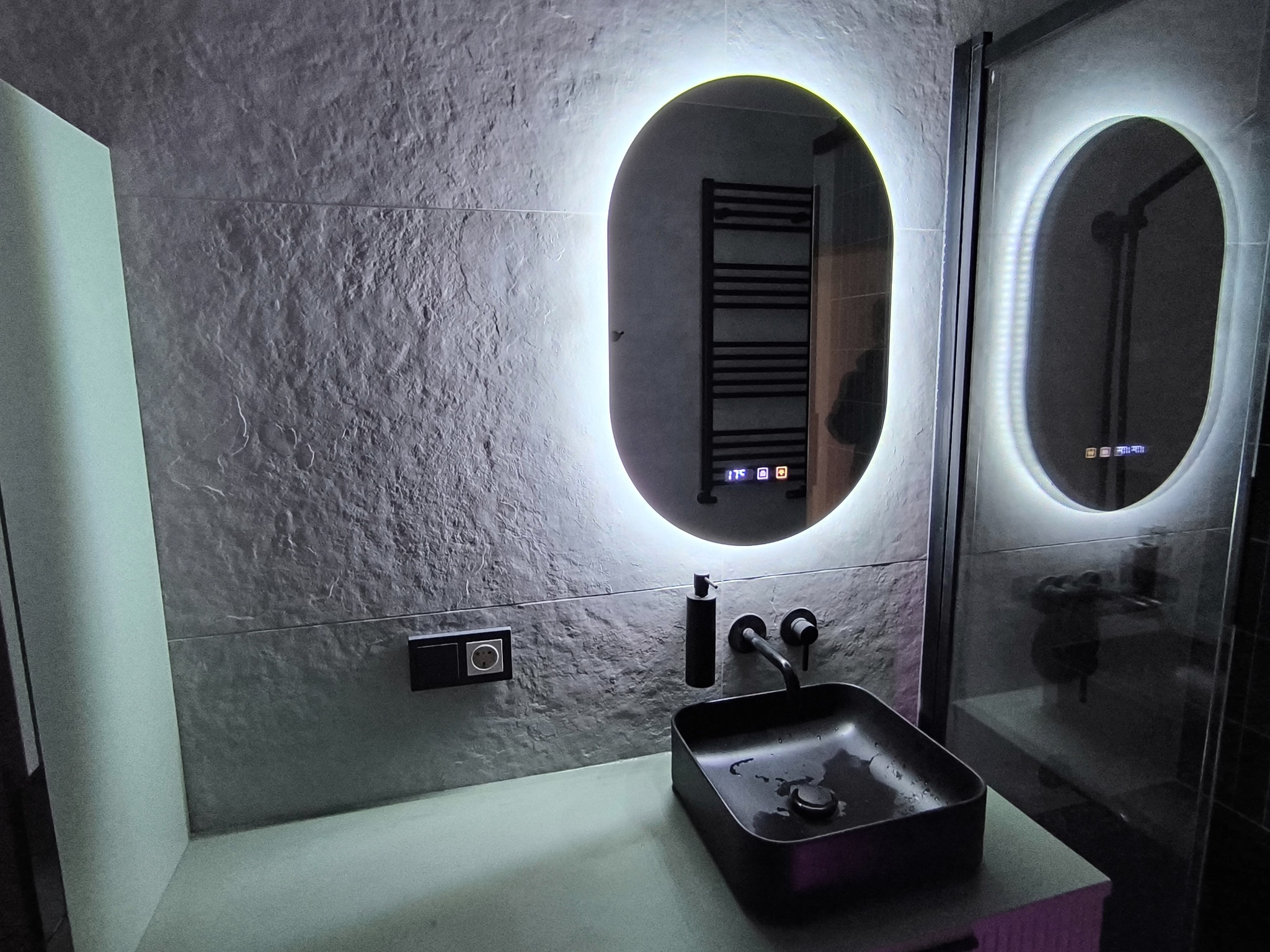 Nowoczesna łazienka z owalnym lustrem LED, czarną umywalką nablatową i szarą, teksturowaną ścianą. Widoczny fragment kabiny prysznicowej z podświetlanym lustrem.