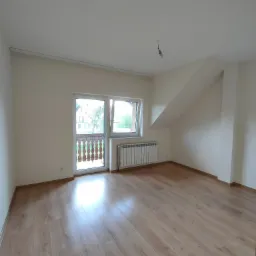 Puste, jasne pomieszczenie na poddaszu z drewnianą podłogą, białymi ścianami i oknem balkonowym, z widocznym grzejnikiem i żarówką zwisającą z sufitu.
