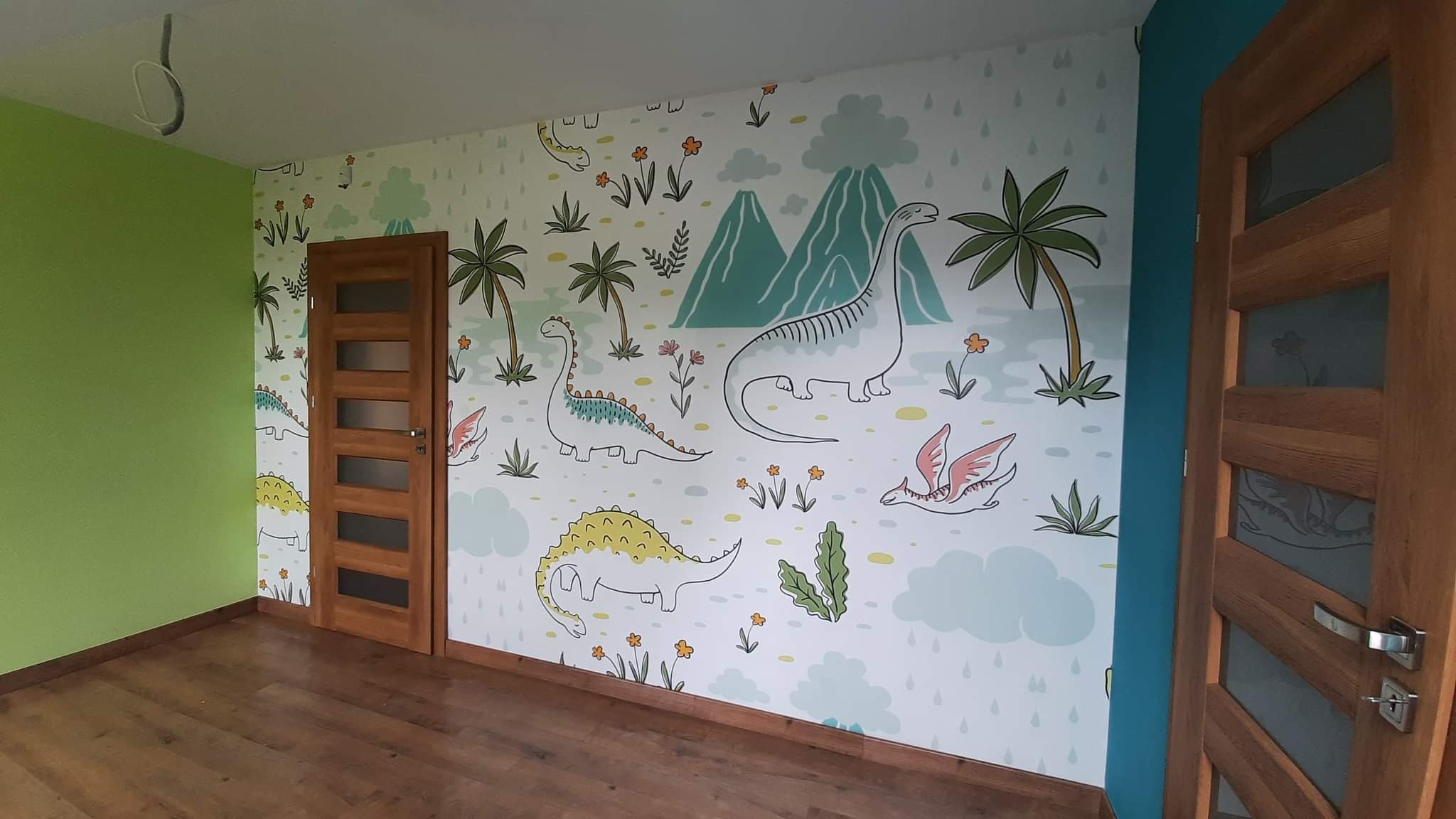 Pokój dziecięcy z zieloną ścianą i ścianą pokrytą tapetą z motywem dinozaurów, widoczne drewniane drzwi z przeszkleniami i podłoga z paneli.