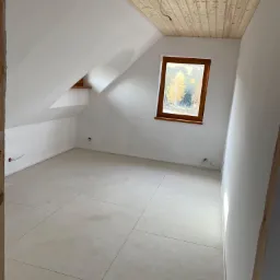 Poddasze z drewnianym sufitem, jasnymi ścianami i podłogą z płytek, okno z widokiem na drzewa.