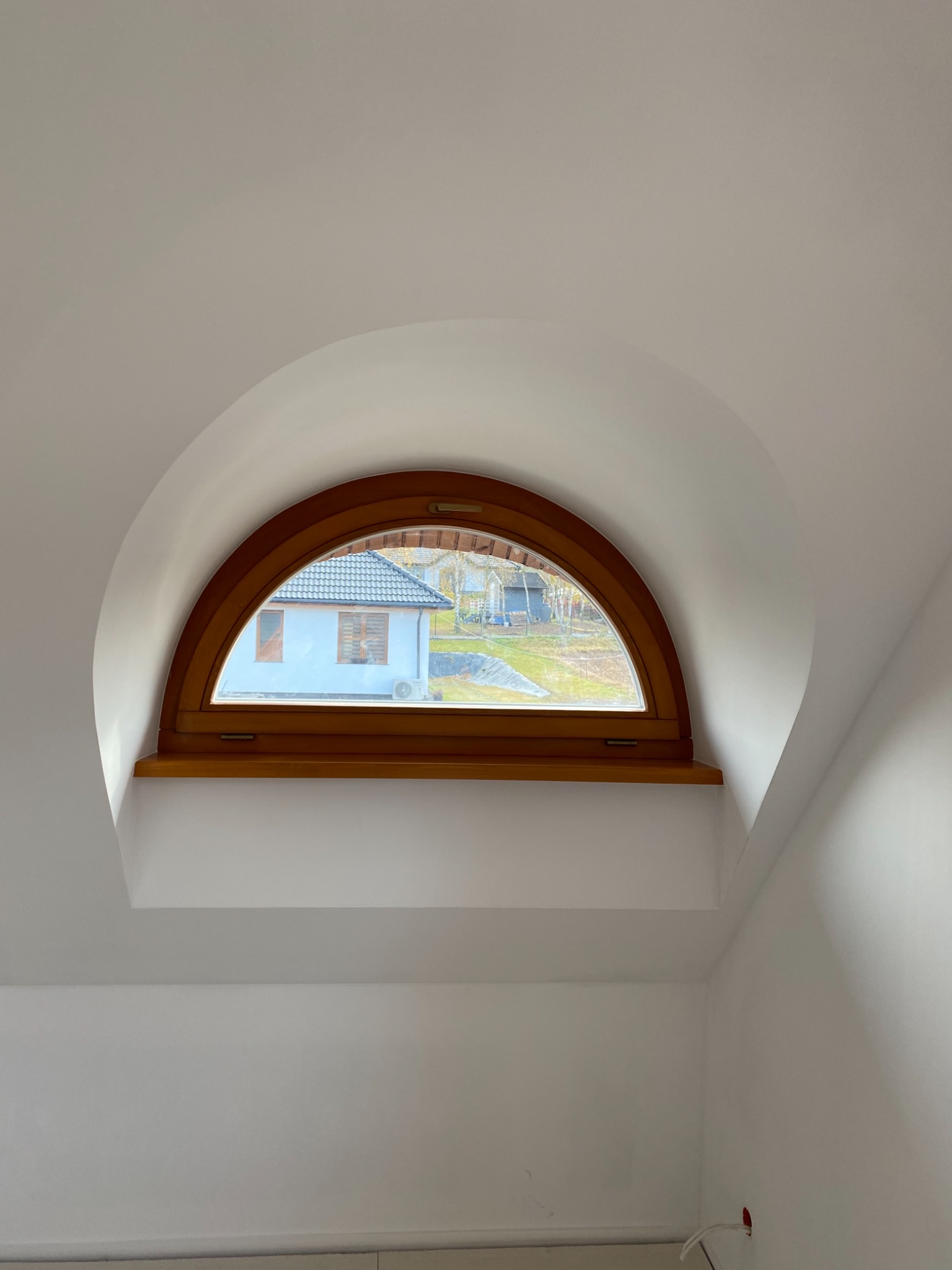 Półokrągłe okno z drewnianą ramą w białym pomieszczeniu na poddaszu, widok na budynek z klimatyzacją na zewnątrz.