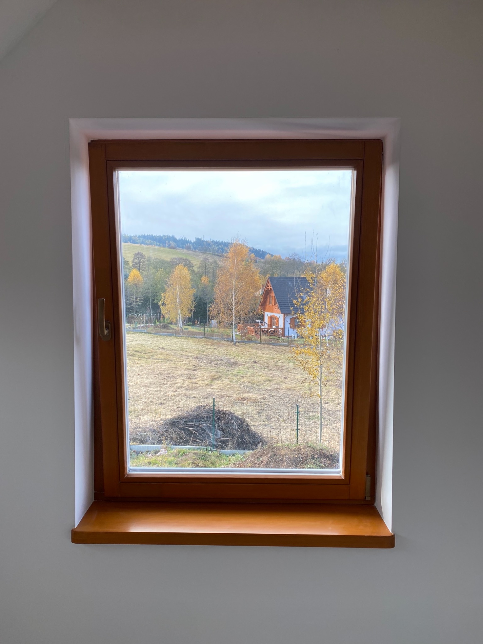 Drewniane okno z widokiem na krajobraz wiejski z domem i drzewami w jesiennej scenerii, widoczne z wnętrza pomieszczenia z białymi ścianami.