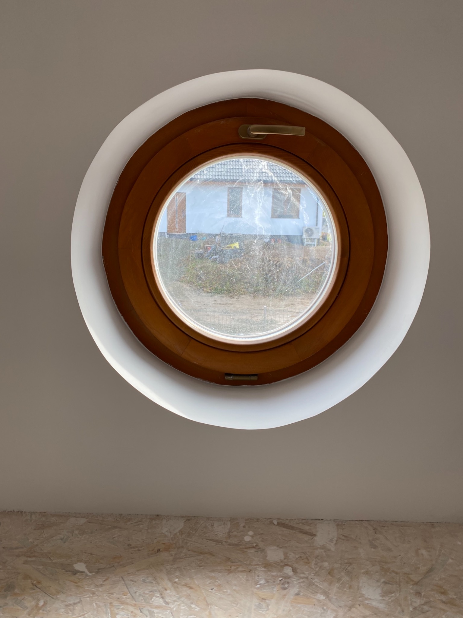 Okrągłe okno z drewnianą ramą i złotą klamką, osadzone w białej ścianie, widok na zewnątrz z zarysami domu i zieleni. Ściana pomalowana na biało, dół ściany wykończony płytą OSB.