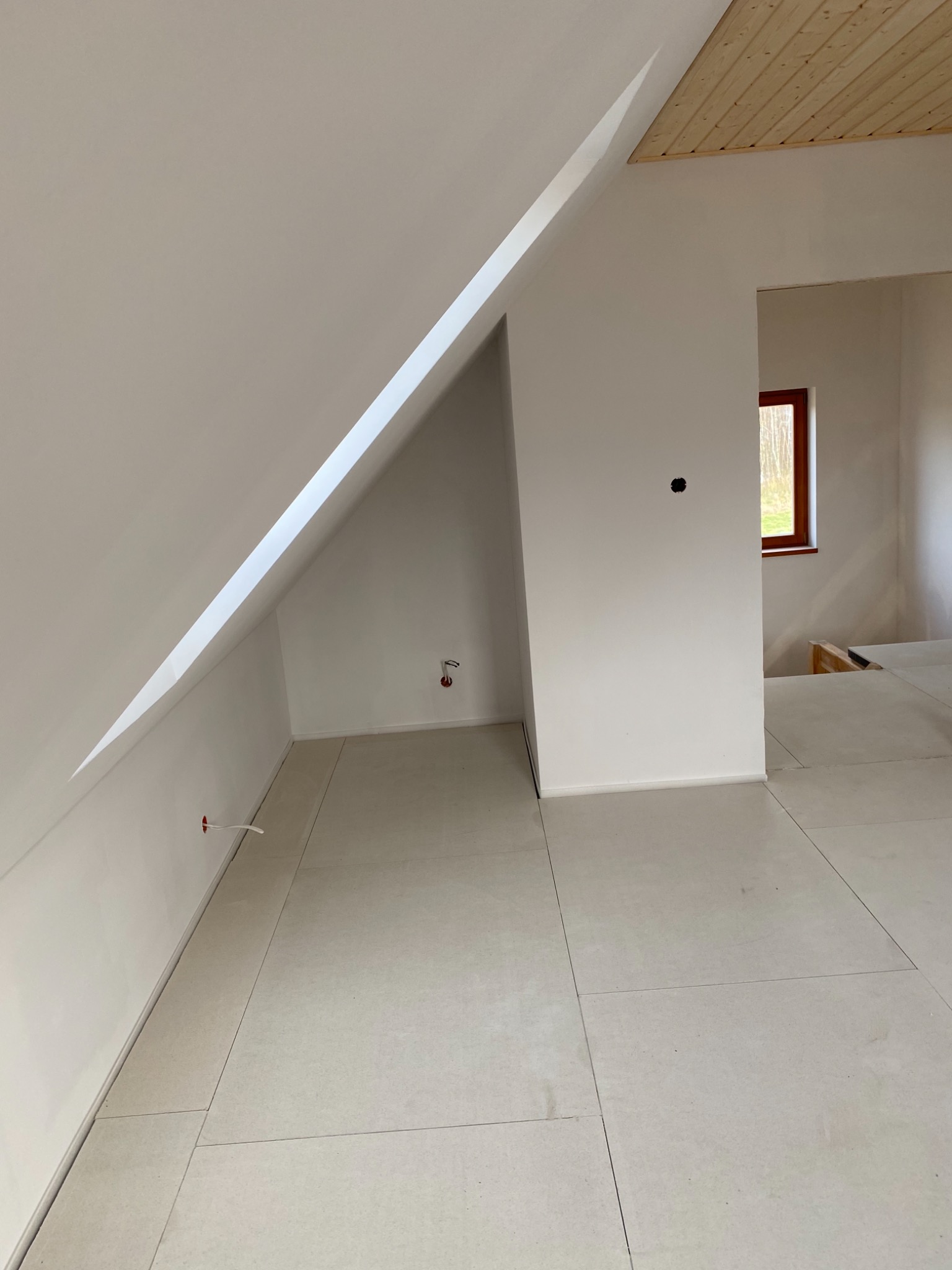 Poddasze po remoncie: białe ściany, ułożona jasna podłoga z płyt, widoczne kable elektryczne wystające ze ściany, drewniane wykończenie sufitu i okno z brązową ramą.