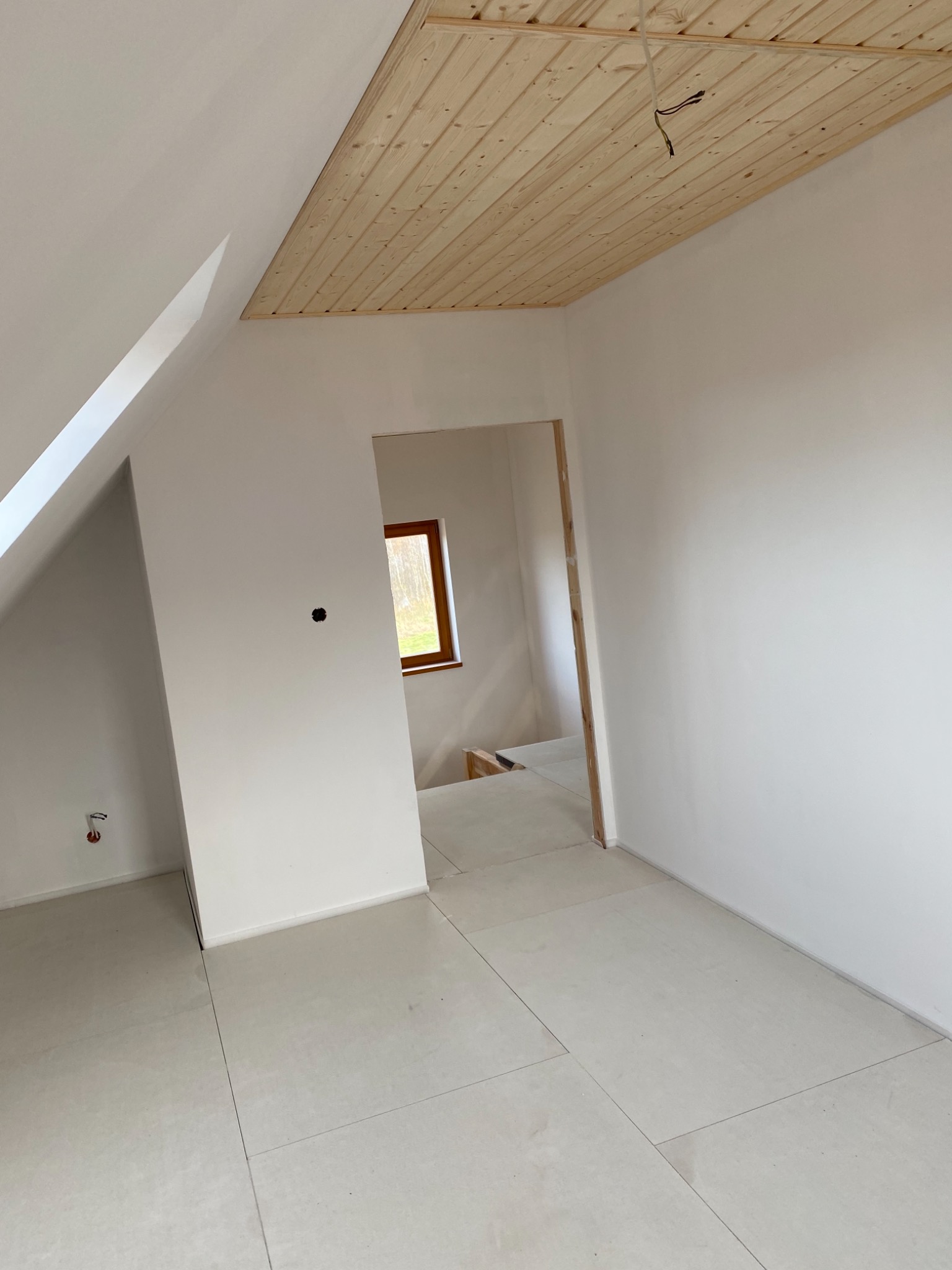 Poddasze w trakcie wykończenia: białe ściany, drewniany sufit z widocznymi kablami, jasna podłoga z płytek, widok na okno w sąsiednim pomieszczeniu przez otwór drzwiowy bez ościeżnicy.