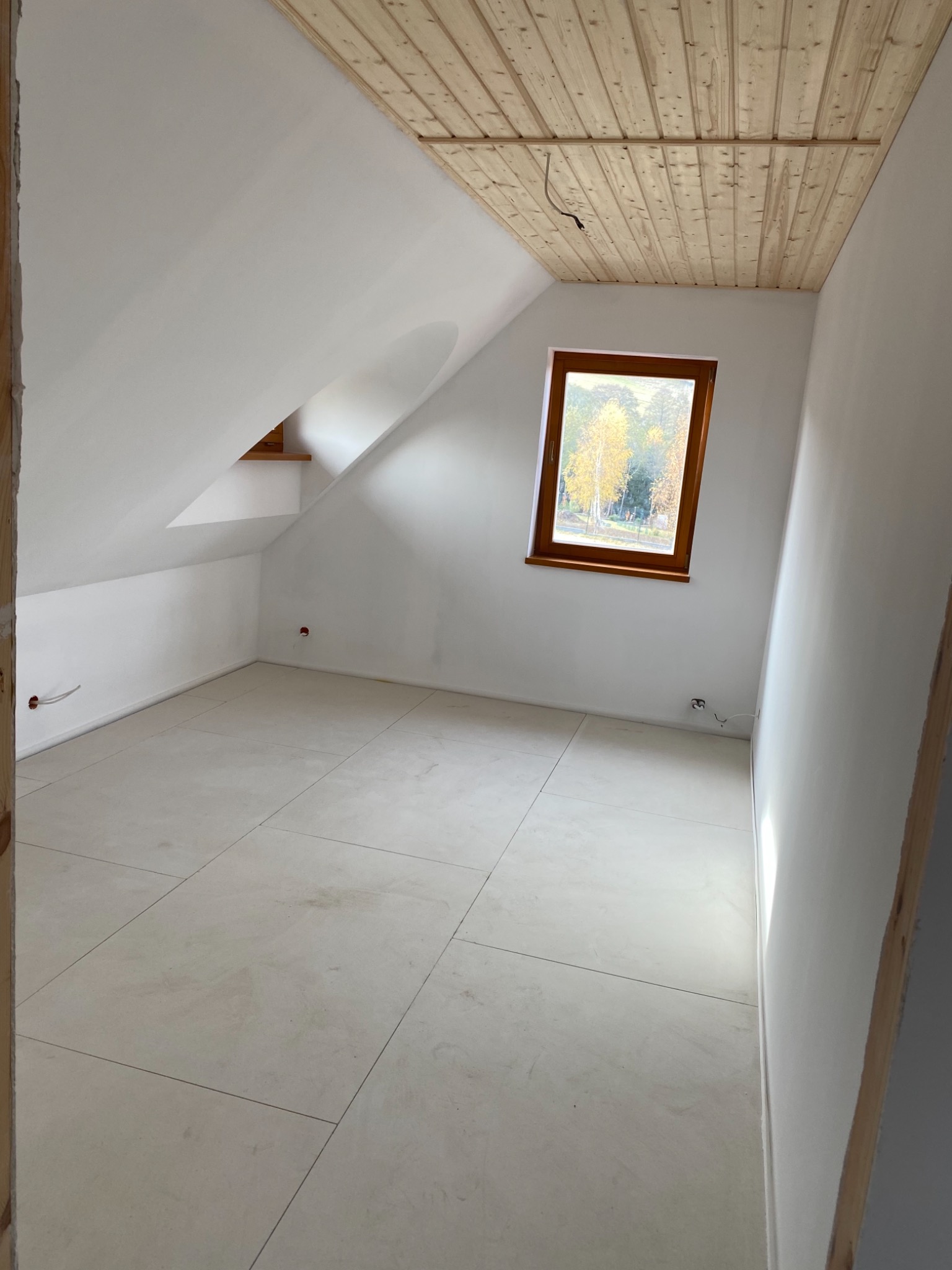 Poddasze z drewnianym sufitem, jasnymi ścianami i podłogą z płytek, okno z widokiem na drzewa.