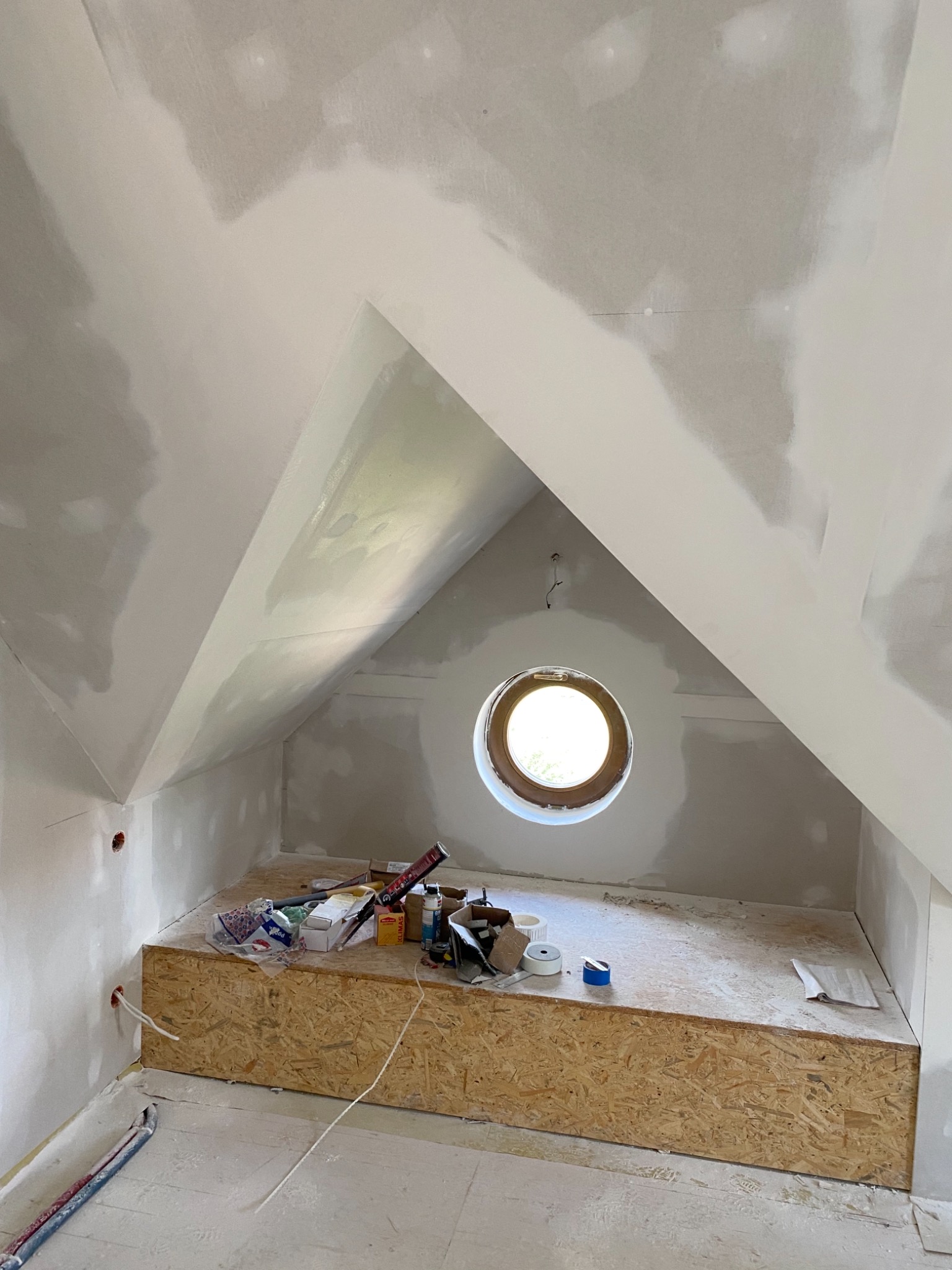 Poddasze w trakcie prac wykończeniowych: ściany i sufit pokryte warstwą szpachli, okrągłe okno w szczycie, widoczne narzędzia i materiały na podwyższeniu z płyty OSB.