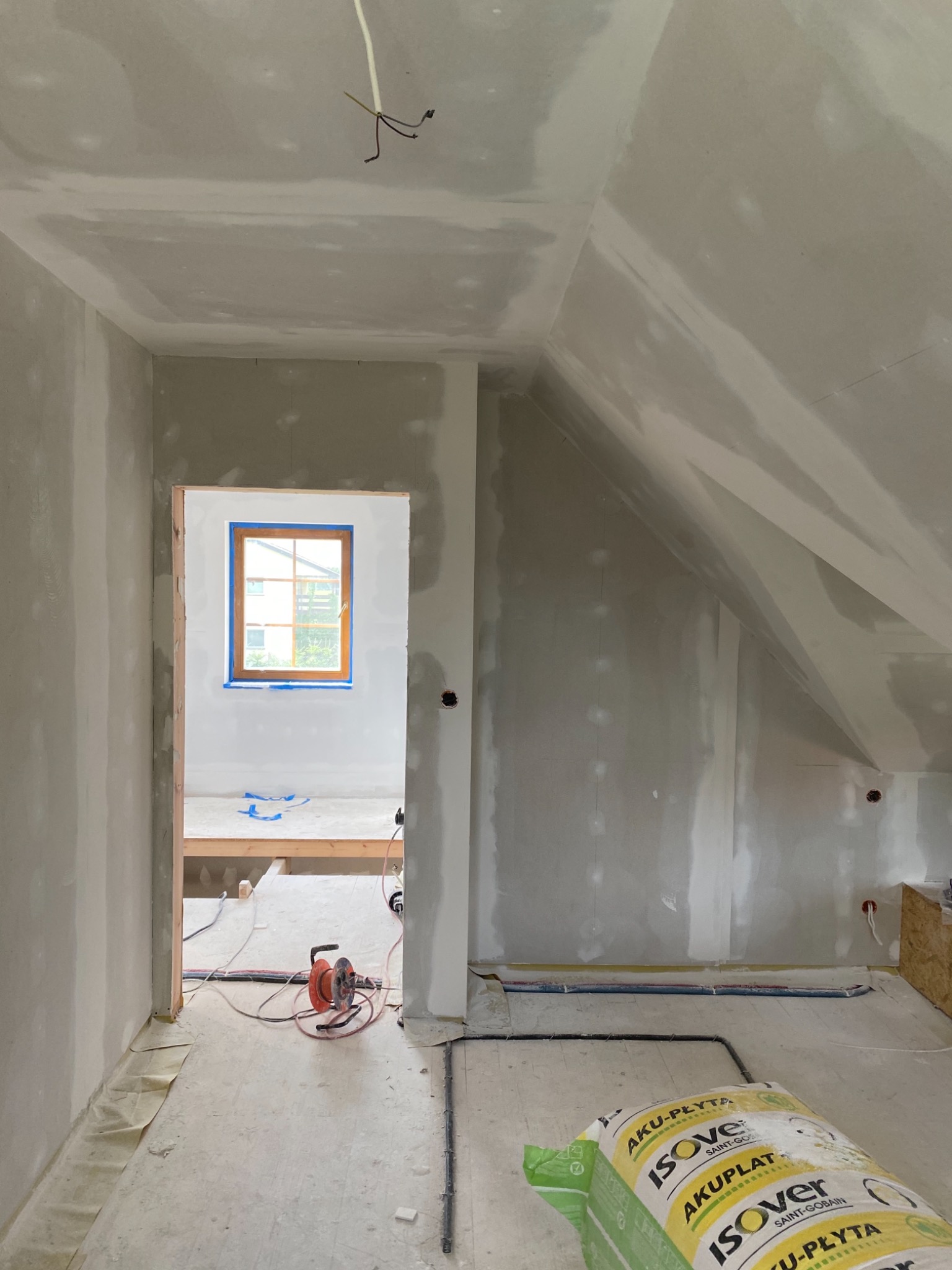 Poddasze w trakcie remontu, ściany pokryte szpachlą na płytach kartonowo-gipsowych, widoczne kable elektryczne, okno zabezpieczone taśmą malarską, na podłodze leży opakowanie płyt izolacyjnych...