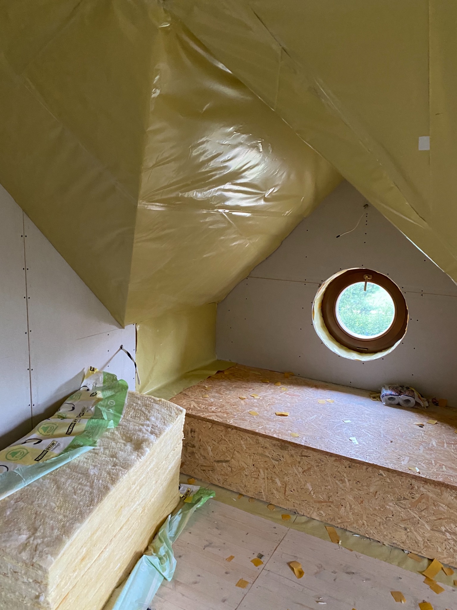Poddasze w trakcie izolacji, widoczne płyty kartonowo-gipsowe, folia paroizolacyjna, okrągłe okno i wełna mineralna w opakowaniu.