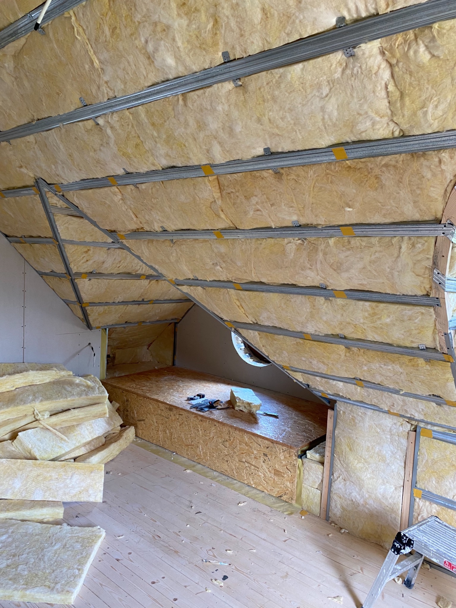 Izolacja poddasza wełną mineralną, widoczne metalowe profile, płyta OSB, drewniana podłoga i fragment drabiny.