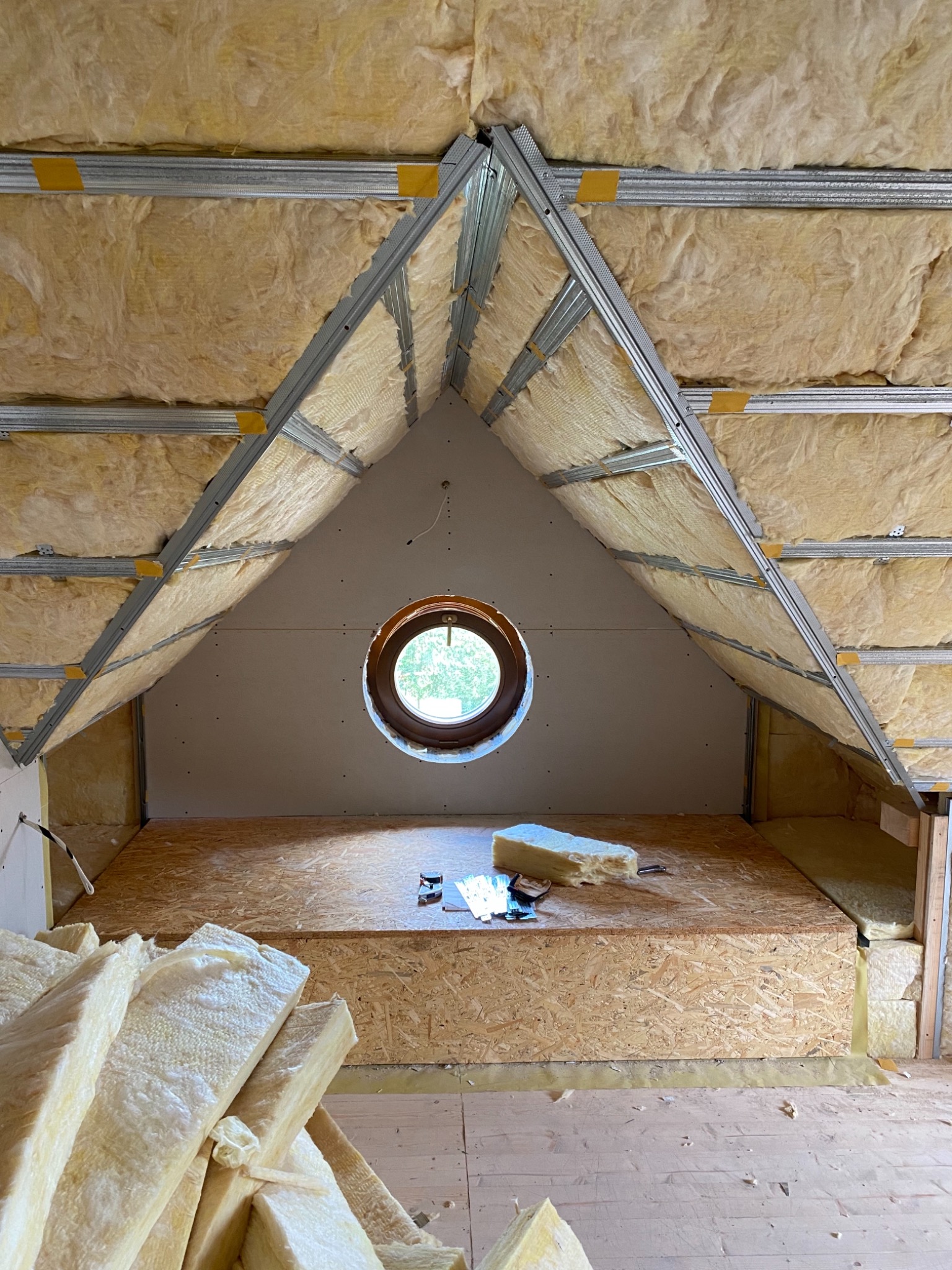 Poddasze w trakcie izolacji: wełna mineralna w metalowej konstrukcji, okrągłe okno w ścianie z płyt kartonowo-gipsowych, płyta OSB na podłodze i narzędzia w tle.