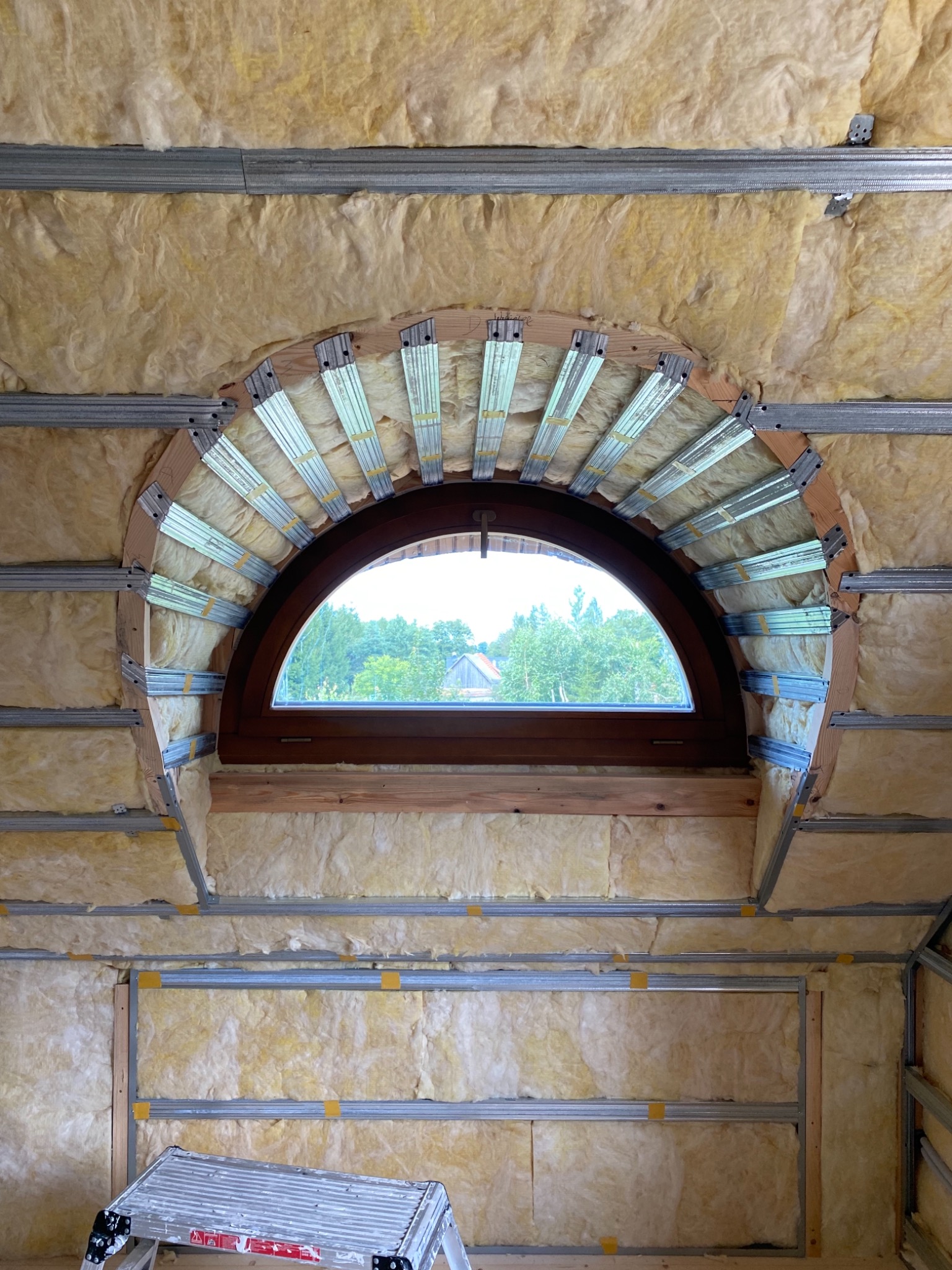 Półokrągłe okno dachowe w trakcie montażu, widoczne ocieplenie wełną mineralną i metalowa konstrukcja szkieletowa, drabina aluminiowa w dolnym rogu.