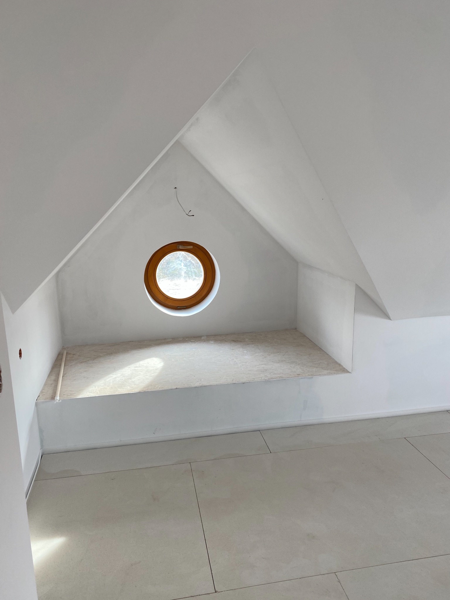 Poddasze z oknem w kształcie bulaja, świeżo po pracach wykończeniowych, ściany i sufit pomalowane na biało, jasne płytki na podłodze, widoczne kable wystające ze ściany.