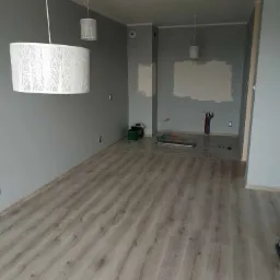 Wnętrze w trakcie remontu: położona podłoga z paneli imitujących drewno, ściany pomalowane na szaro, widoczne przygotowania do prac wykończeniowych na ścianie, wiszące lampy z ażurowym wzorem.