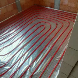 Czerwone rurki ogrzewania podłogowego ułożone spiralnie na folii refleksyjnej z siatką, przymocowane czarnymi klipsami, widoczne surowe mury z cegły i betonowy słup w tle.