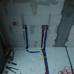 Surowe wnętrze w trakcie prac instalacyjnych: widoczne rury doprowadzające wodę, narzędzia hydrauliczne rozłożone na podłodze z izolacją, surowe ściany z betonu komórkowego.