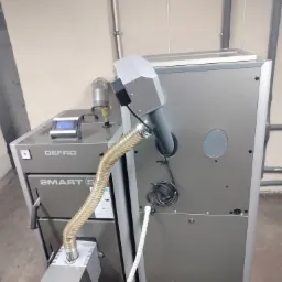 Szary kocioł na paliwo stałe marki DEFRO Smart E z podajnikiem, widoczny w kotłowni na tle beżowych ścian i szarych rur.