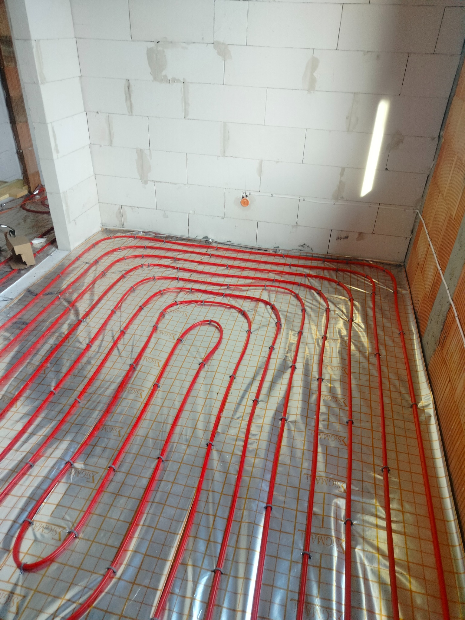 Czerwone rury ogrzewania podłogowego ułożone na folii z siatką, widoczne ściany z białego betonu komórkowego w surowym stanie, w tle fragment instalacji.