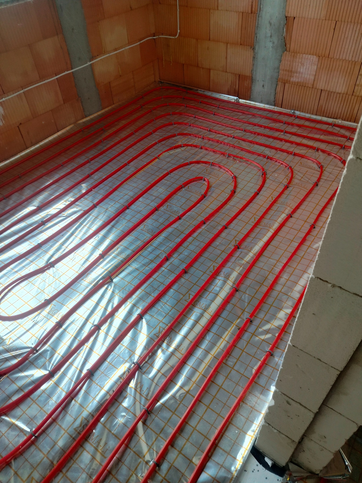 Czerwone rurki ogrzewania podłogowego ułożone spiralnie na folii refleksyjnej z siatką, przymocowane czarnymi klipsami, widoczne surowe mury z cegły i betonowy słup w tle.