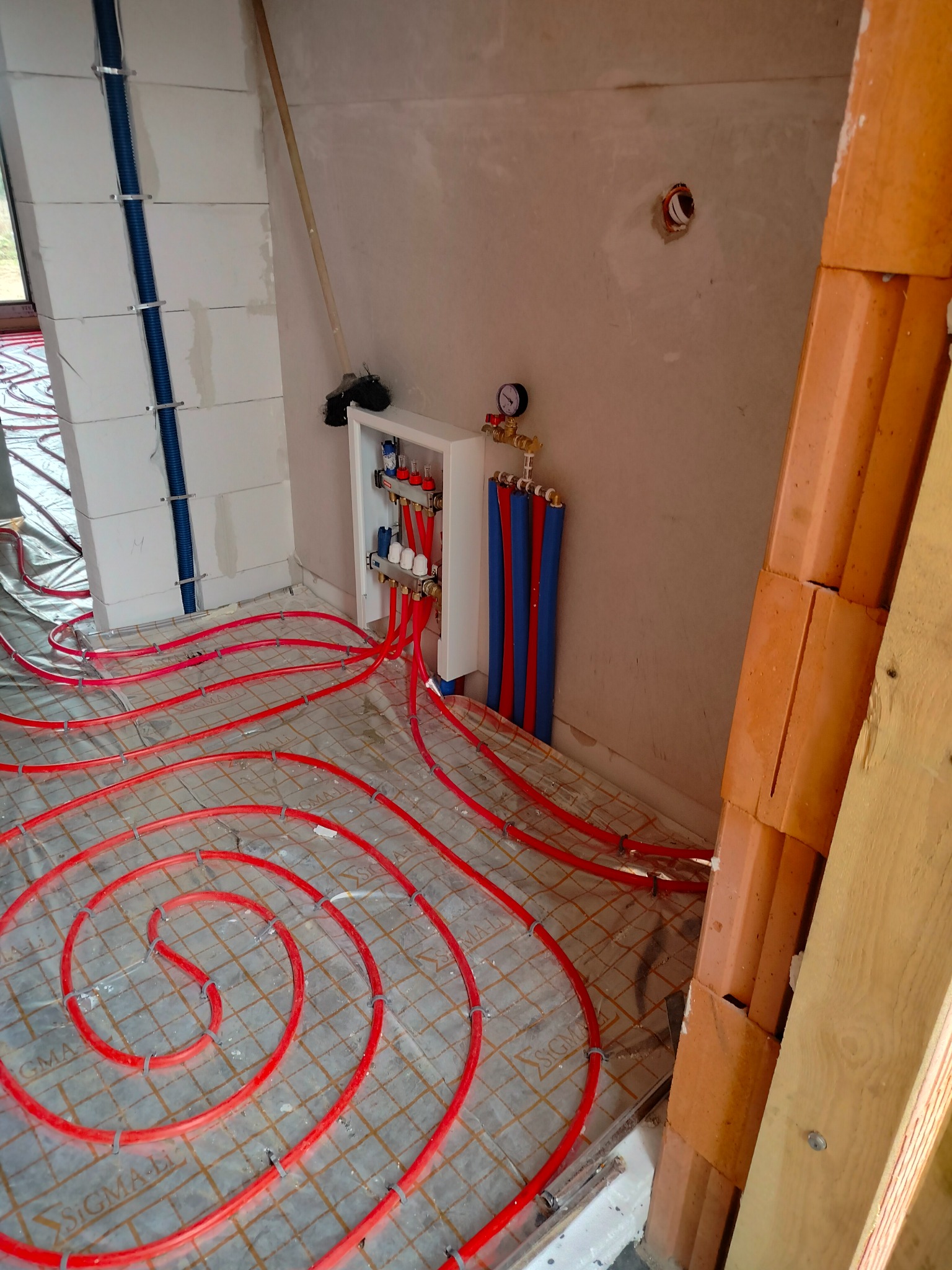 Rozłożone czerwone rury ogrzewania podłogowego na folii z siatką, widoczny rozdzielacz w białej szafce z manometrem, ściany w surowym stanie, w tle bloczki z niebieską rurą oraz stojąca miotła.