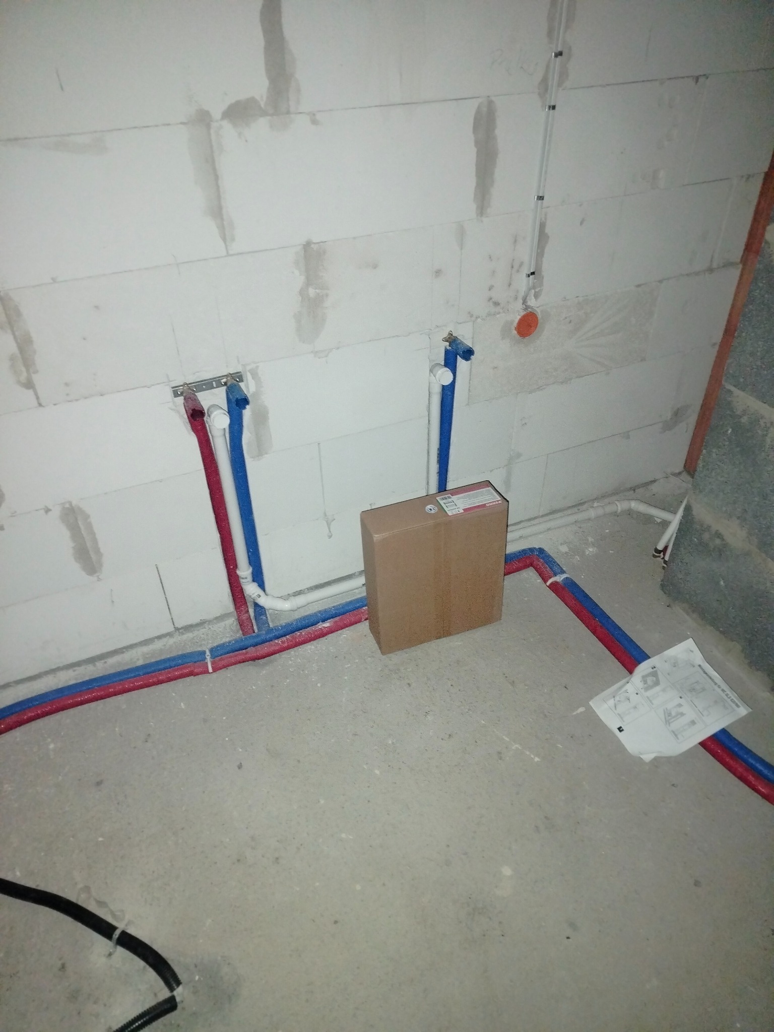 Surowy stan: Instalacja wodna z rur PEX czerwonych i niebieskich, wystających ze ściany z betonu komórkowego, połączonych na podłodze, z kartonem i instrukcją obok.