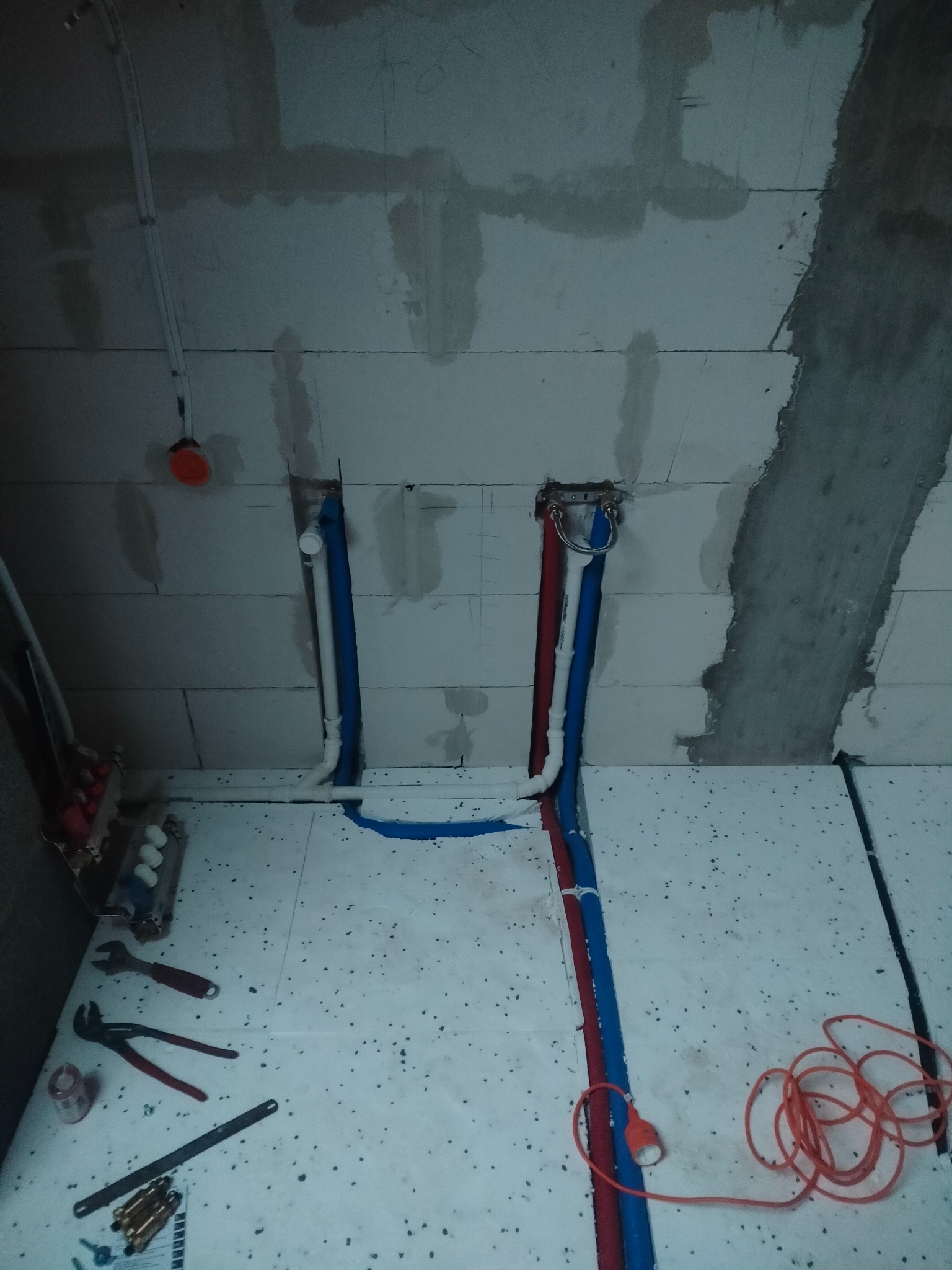 Surowe wnętrze w trakcie prac instalacyjnych: widoczne rury doprowadzające wodę, narzędzia hydrauliczne rozłożone na podłodze z izolacją, surowe ściany z betonu komórkowego.