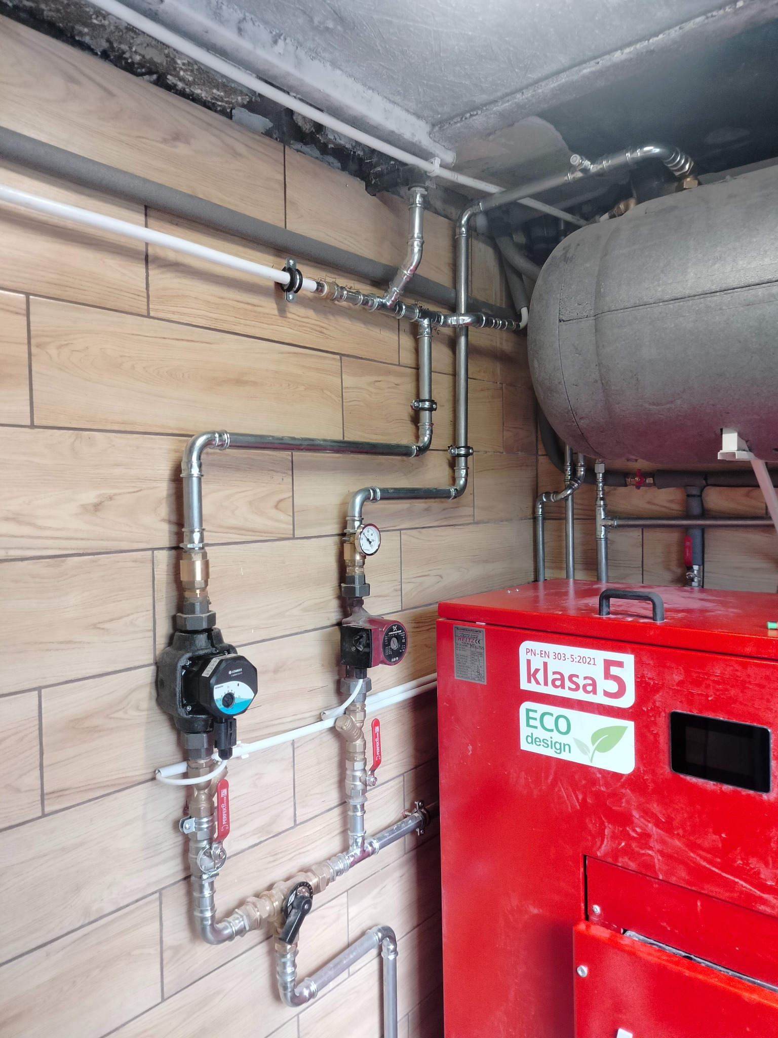 Czerwony piec grzewczy z instalacją hydrauliczną i zbiornikiem wyrównawczym na tle ściany wyłożonej drewnopodobnymi panelami.