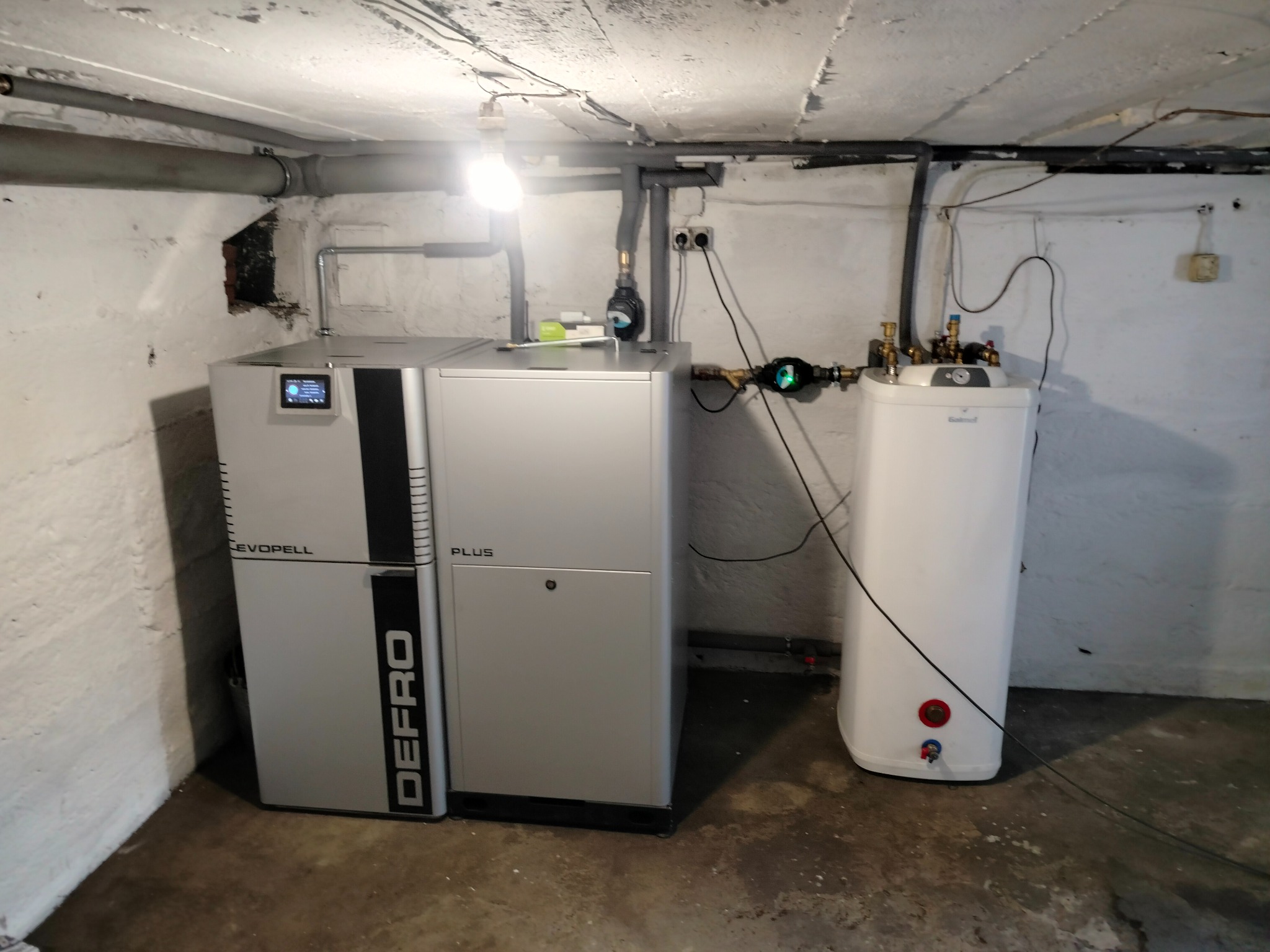 Nowoczesny piec na pellet marki Defro Evopell Plus w kotłowni, obok biały zasobnik ciepłej wody użytkowej, widoczne rury i instalacja.