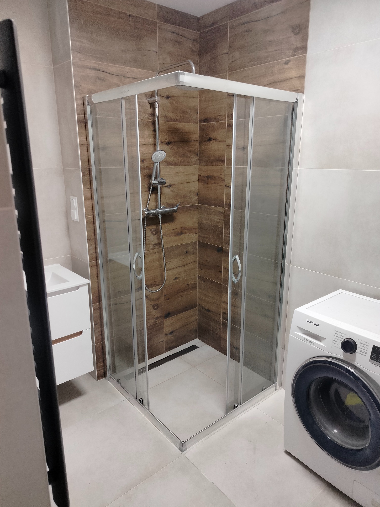 Nowoczesna kabina prysznicowa z przesuwnymi drzwiami, chromowanymi profilami i deszczownicą, w łazience wykończonej płytkami imitującymi drewno i jasnymi płytkami podłogowymi, obok pralka Samsung.