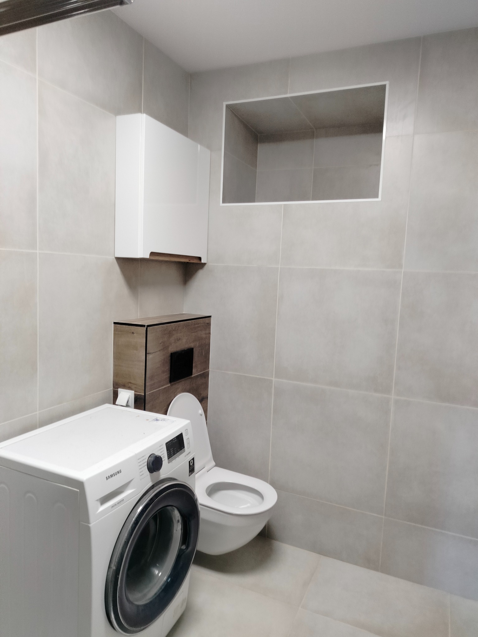 Jasna łazienka po remoncie w Pucku: pralka Samsung, podwieszana toaleta z zabudową drewnopodobną, szafka wisząca i wnęka w płytkach.