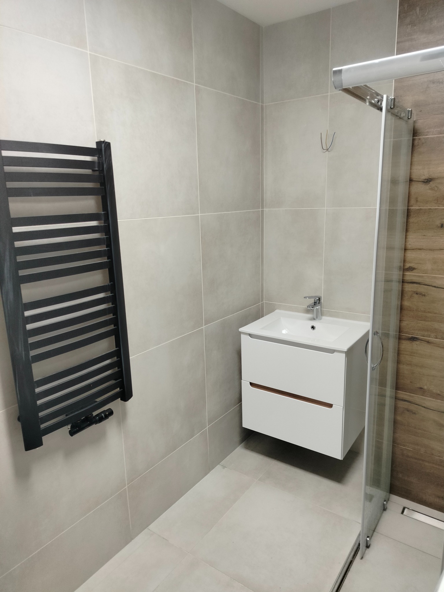 Nowoczesna łazienka z szarymi płytkami, grzejnikiem drabinkowym, białą szafką umywalkową z drewnianymi akcentami i kabiną prysznicową z drewnopodobnymi panelami na ścianie.