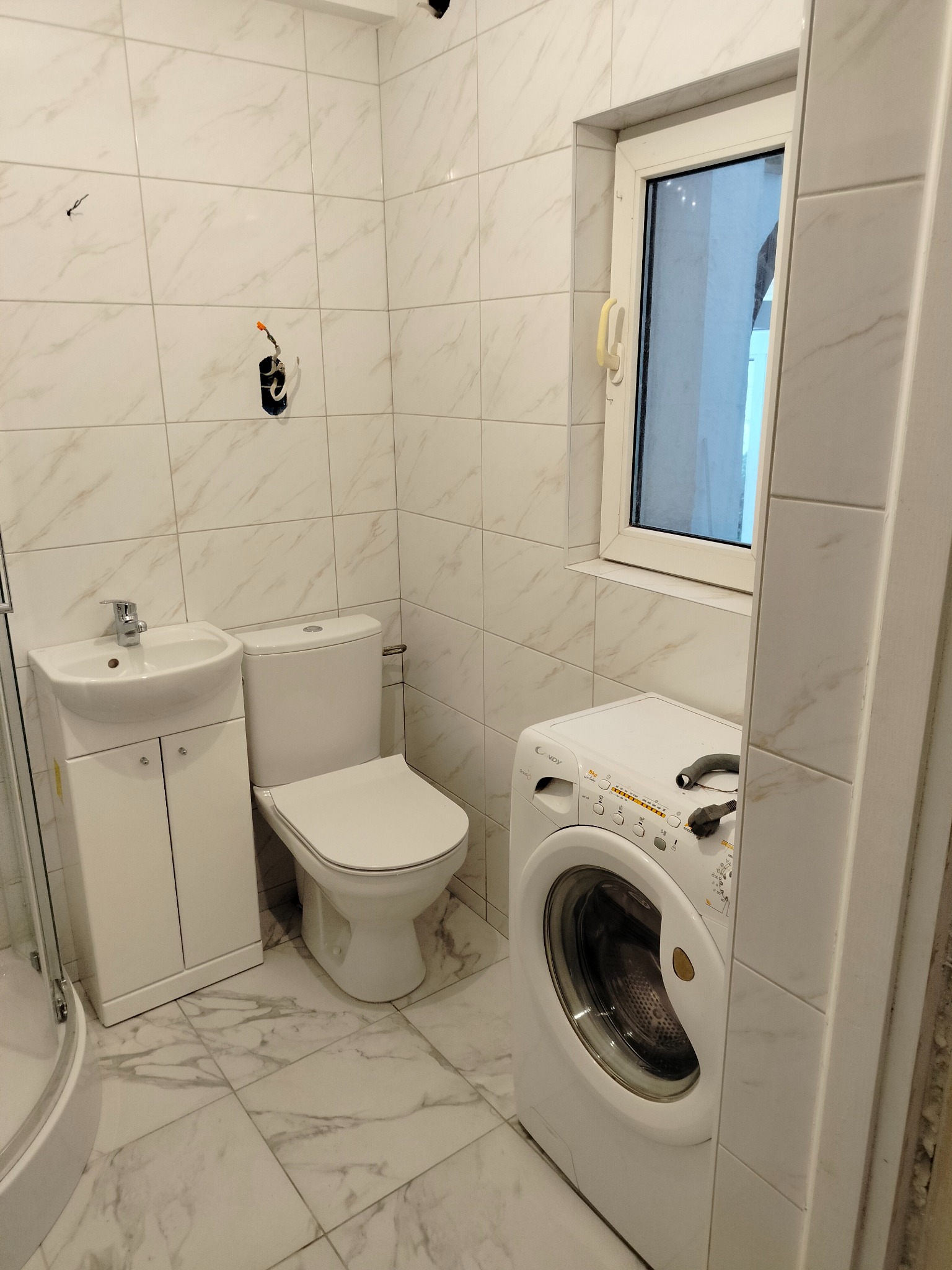 Jasna łazienka w trakcie remontu: białe marmurowe płytki, umywalka z szafką, toaleta, pralka Candy i niedokończone podłączenie elektryczne na ścianie.