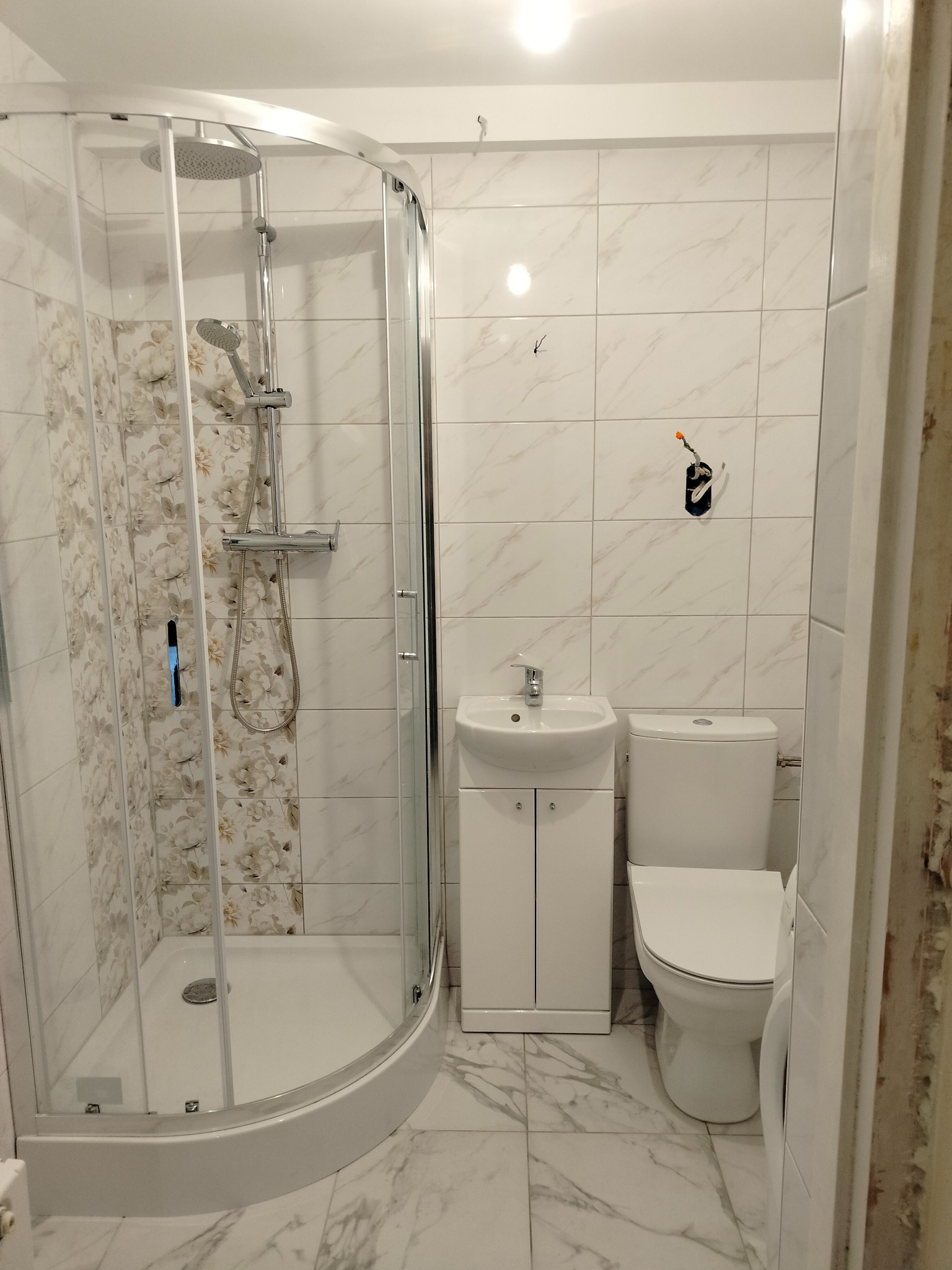 Nowoczesna łazienka po remoncie: kabina prysznicowa z motywem kwiatowym, umywalka z szafką i toaleta na marmurowej podłodze, widoczne drobne niedociągnięcia wykończeniowe.