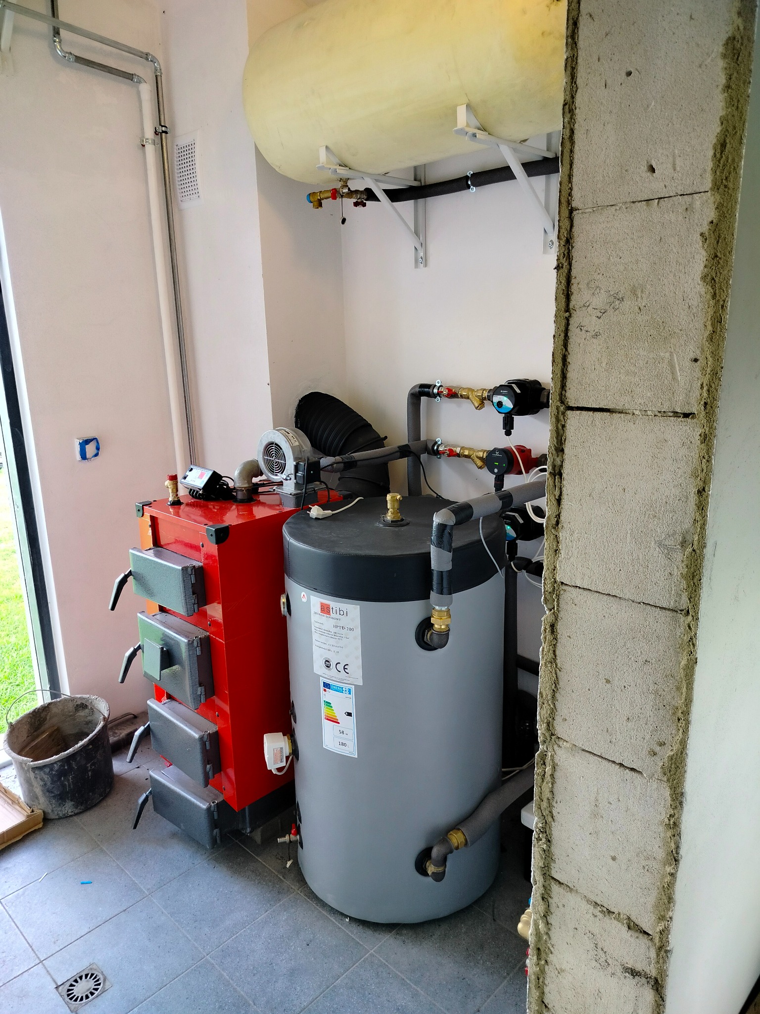 Instalacja kotła na paliwo stałe z zasobnikiem ciepłej wody użytkowej w kotłowni, widoczny wentylator nadmuchowy, rury instalacyjne oraz zbiornik wyrównawczy pod sufitem.