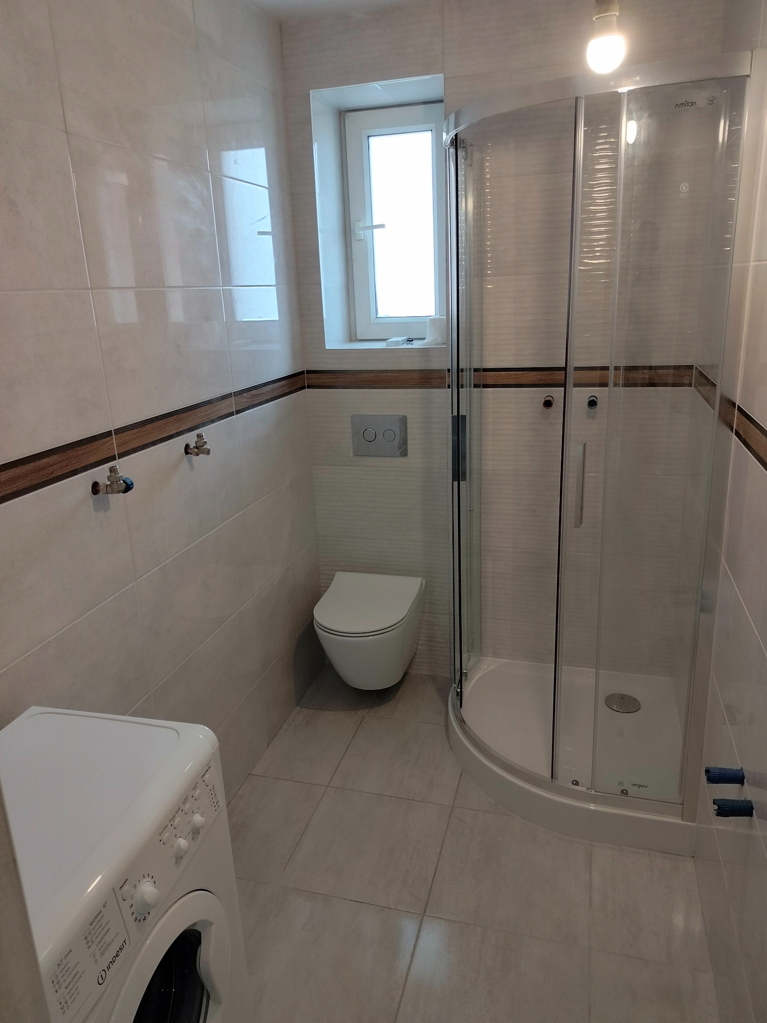 Jasna łazienka po remoncie z kabiną prysznicową, toaletą podwieszaną i pralką Indesit, wykończona płytkami z dekoracyjnym pasem.