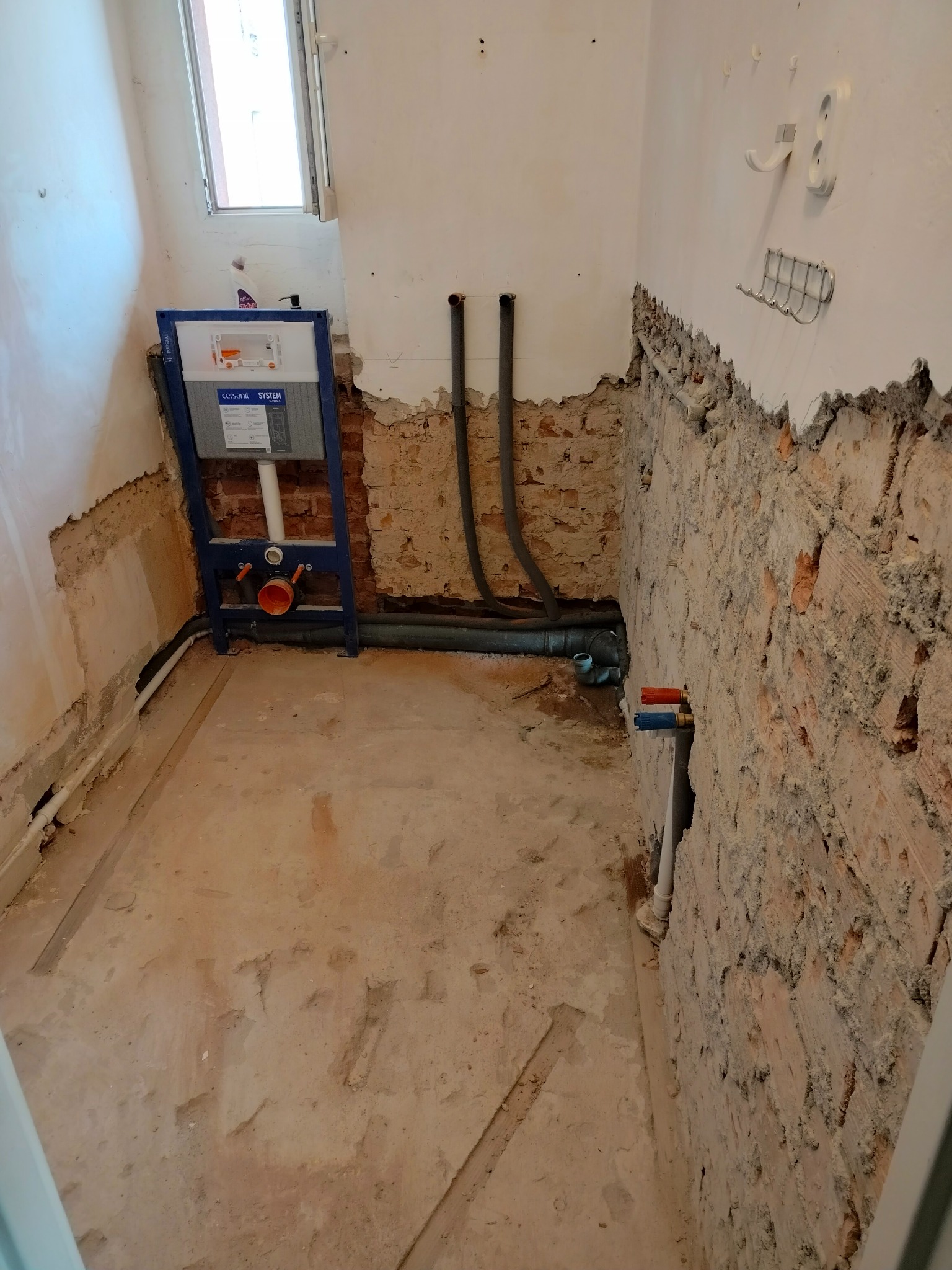 Surowy stan łazienki w trakcie remontu: odsłonięta cegła, instalacja podtynkowa Cersanit i rury kanalizacyjne.