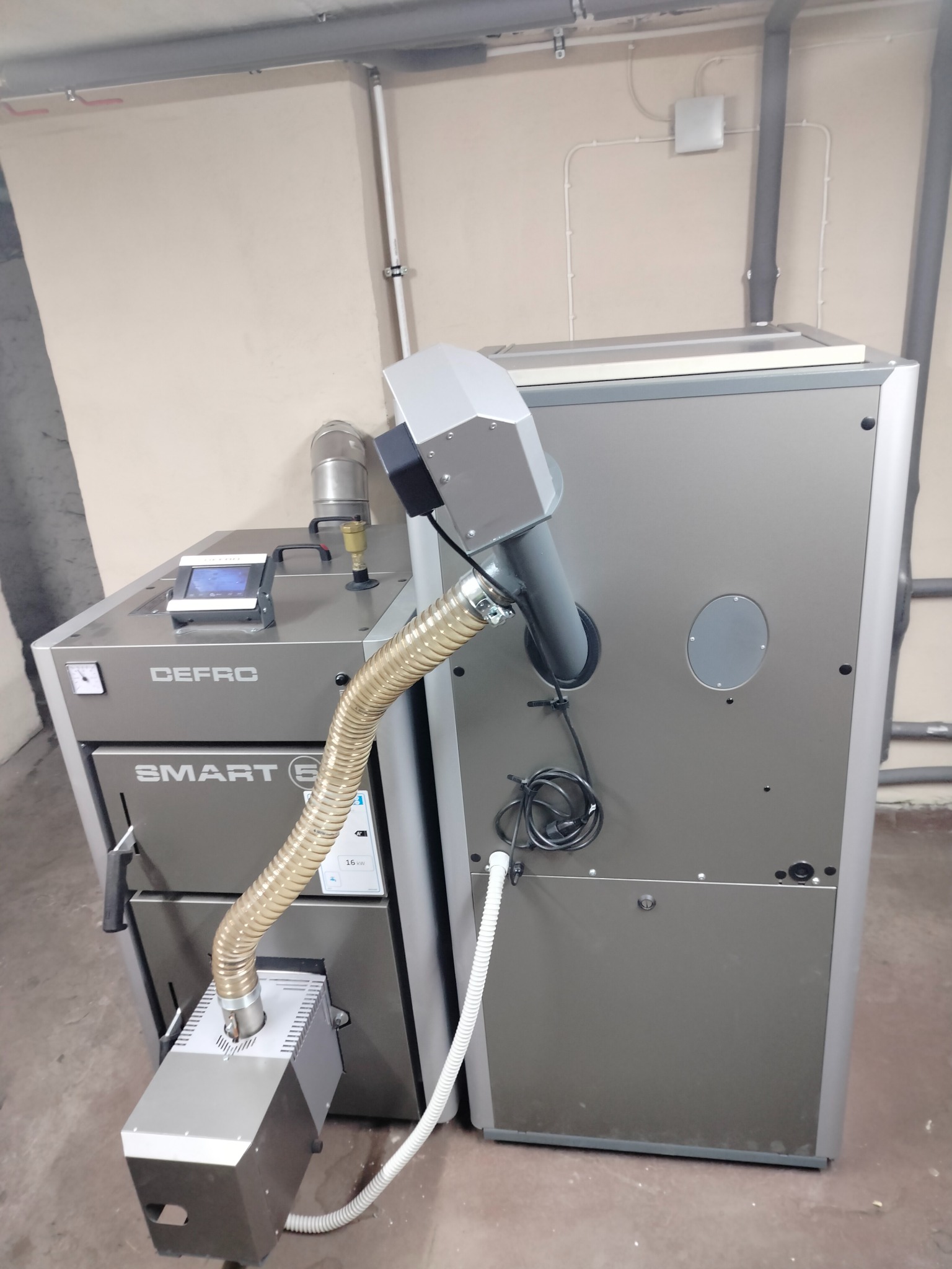 Szary kocioł na paliwo stałe marki DEFRO Smart E z podajnikiem, widoczny w kotłowni na tle beżowych ścian i szarych rur.