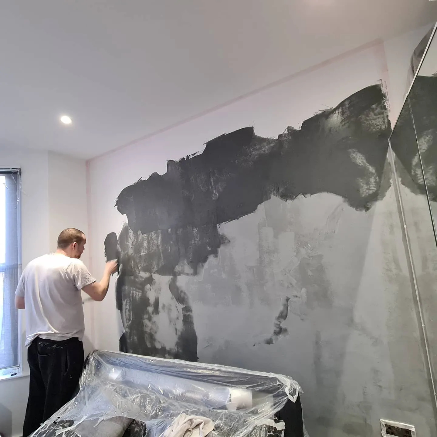 Mężczyzna w białej koszulce nakłada szarą masę szpachlową na ścianę w pokoju, widoczny fragment okna i meble zabezpieczone folią.