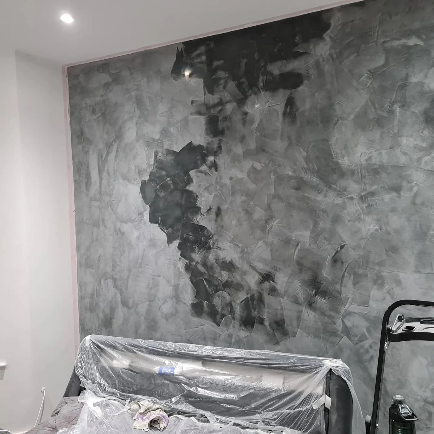 Ściana w trakcie malowania dekoracyjną farbą strukturalną w odcieniach szarości, częściowo pokryta świeżą warstwą, z zabezpieczonym folią meblem i fragmentem drabiny malarskiej w kadrze.