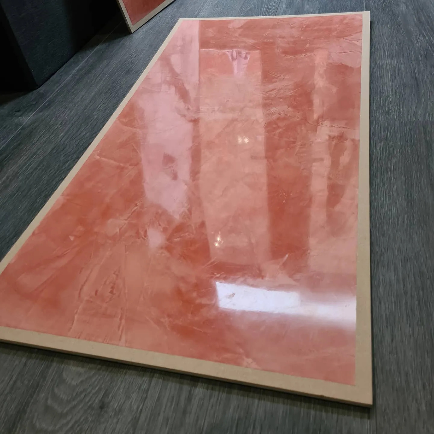 Próbka dekoracyjnej, marmurkowej powłoki w odcieniach różu i beżu, umieszczona na drewnianej płycie, leżąca na szarych panelach podłogowych.