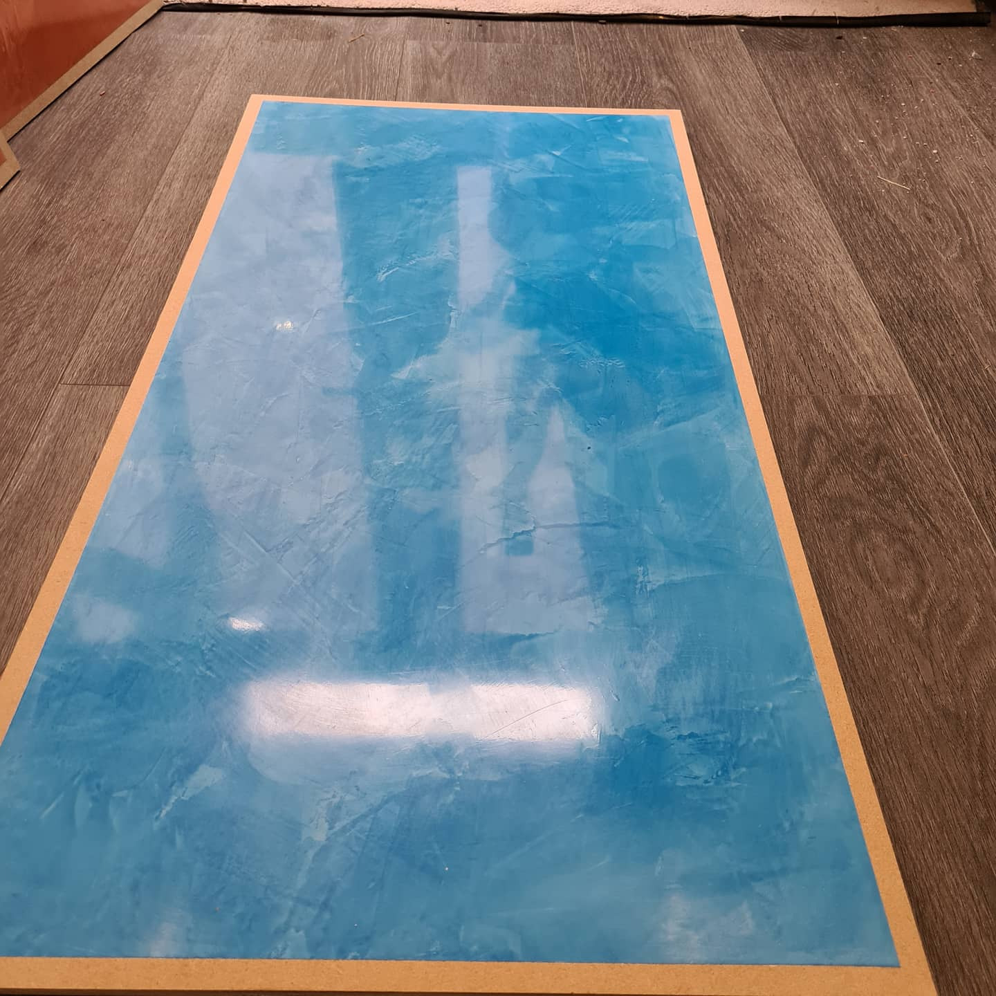 Niebieska, błyszcząca powierzchnia z efektem marmuru, otoczona drewnianą ramą na tle podłogi z paneli imitujących drewno.