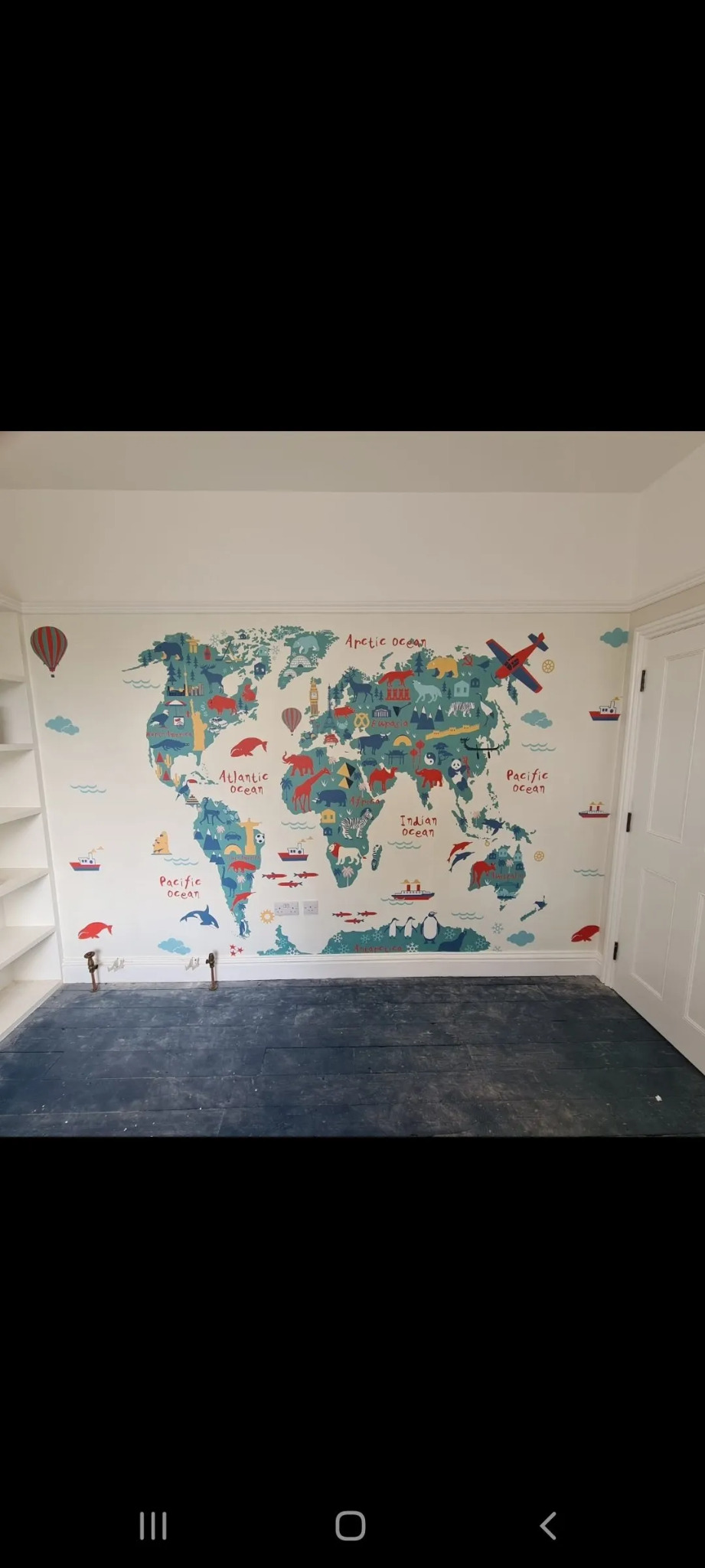Ściana pokoju dziecięcego z naklejoną mapą świata w stylu kreskówkowym, przedstawiającą zwierzęta, budynki i pojazdy, z widocznymi wystającymi rurami grzewczymi przy podłodze.