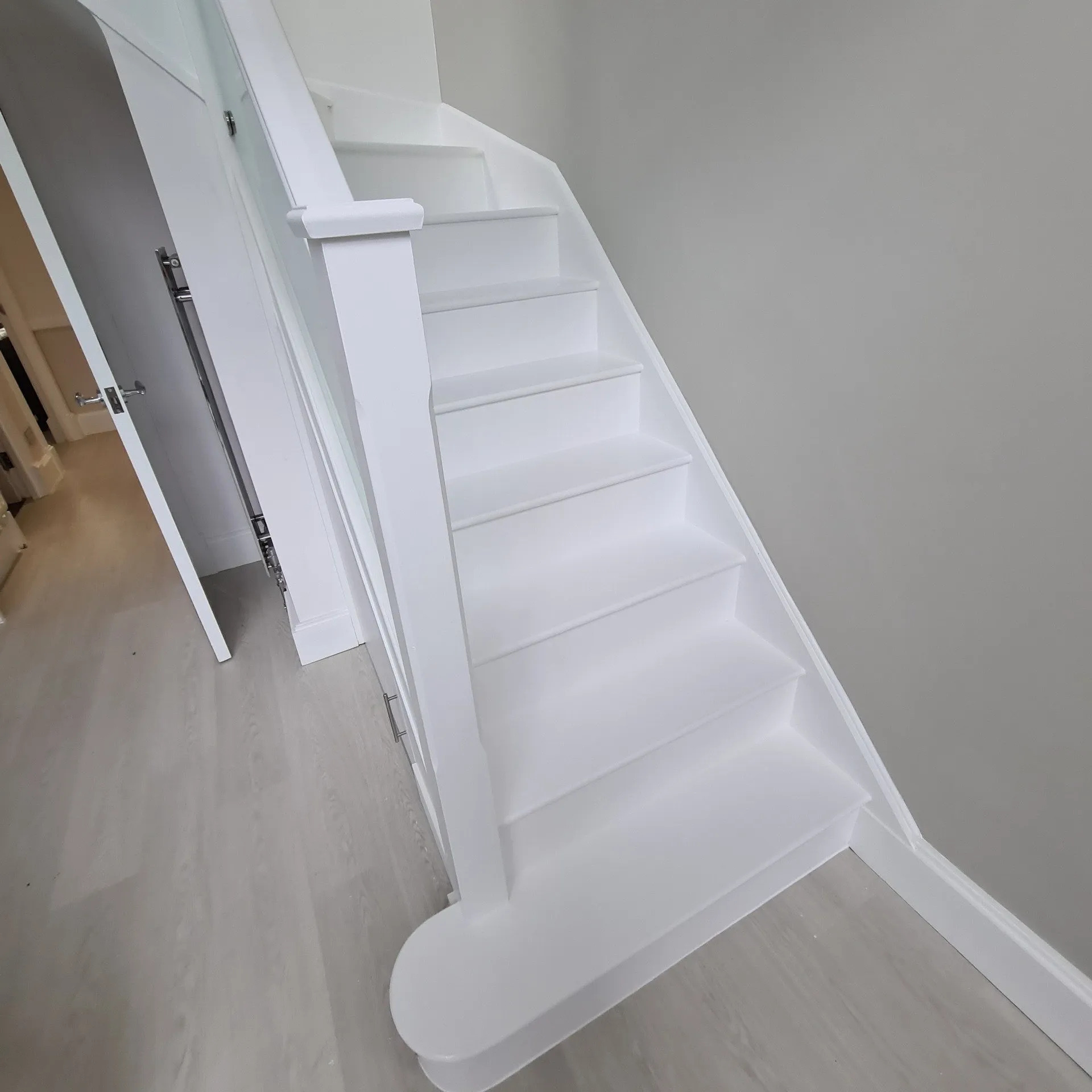 Białe, świeżo odnowione schody z drewnianymi stopniami i barierką, widok z dołu, jasne pomieszczenie z szarą ścianą w tle.