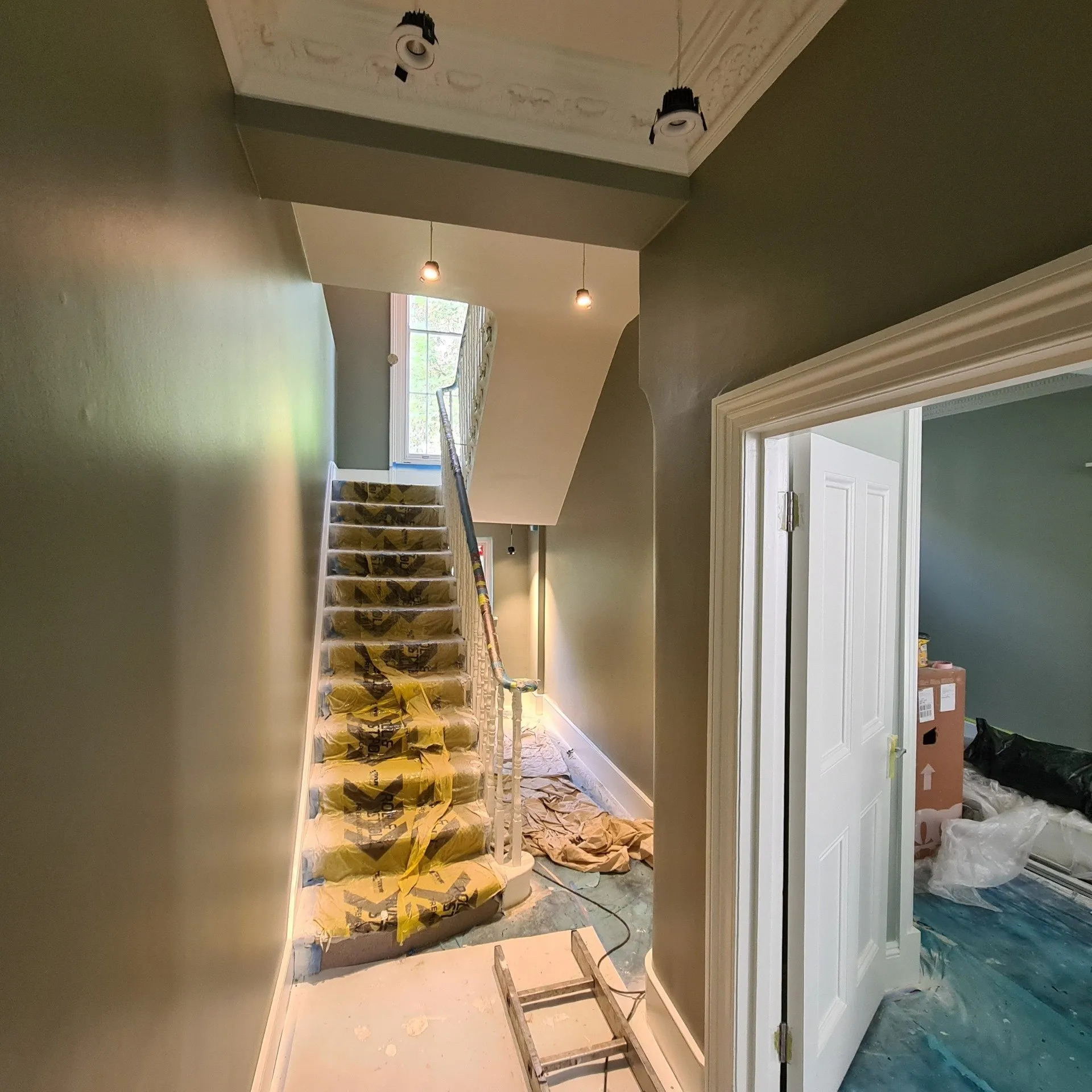 Korytarz i schody w trakcie prac wykończeniowych, zabezpieczone folią malarską, widoczne świeżo pomalowane ściany w odcieniach szarości i bieli, drabina malarska na podłodze.