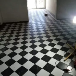 Nowa podłoga w szachownicę z czarno-białych płytek w jasnym pomieszczeniu, widoczna lampa budowlana i wtyczka na podłodze.