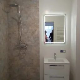 Wykończona łazienka z kabiną prysznicową wyłożoną płytkami imitującymi kamień, białą szafką z umywalką i podświetlanym lustrem, w którym odbijają się dwie osoby.