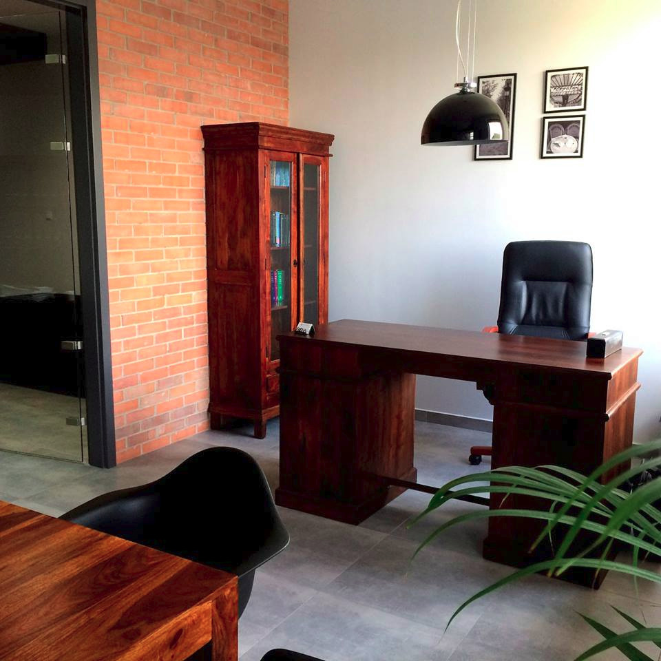 Wnętrze gabinetu z biurkiem z ciemnego drewna, regałem na książki, czarnym krzesłem biurowym i ścianą z cegły, oświetlone wiszącą lampą.