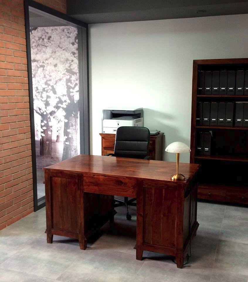 Rustykalne biuro z mahoniowym biurkiem, skórzanym krzesłem i regałem na segregatory, oświetlone lampką z mlecznym kloszem, z widokiem na kwitnące drzewa za oknem.